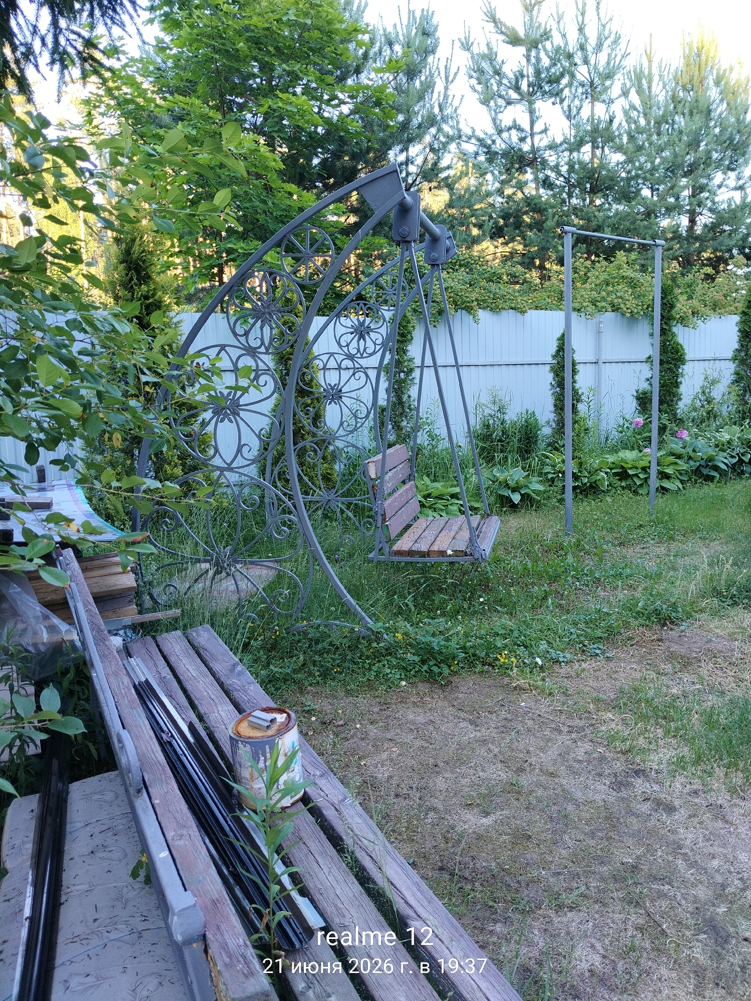 In the city of Космос-2 Снт, Russia, a garden scene unfolds under what appears to be daylight. A metal swing set with a wooden seat stands prominently in the center of the frame, its ornate scrollwork suggesting a decorative design. To the left, a wooden bench with various metal objects and a can of paint rests upon it, hinting at ongoing projects or stored materials. The background is dominated by lush greenery, including trees and a white fence that delineates the property. The ground is covered in grass and patches of dry earth, with scattered wildflowers adding touches of color. The overall impression is one of a lived-in, slightly untidy outdoor space.