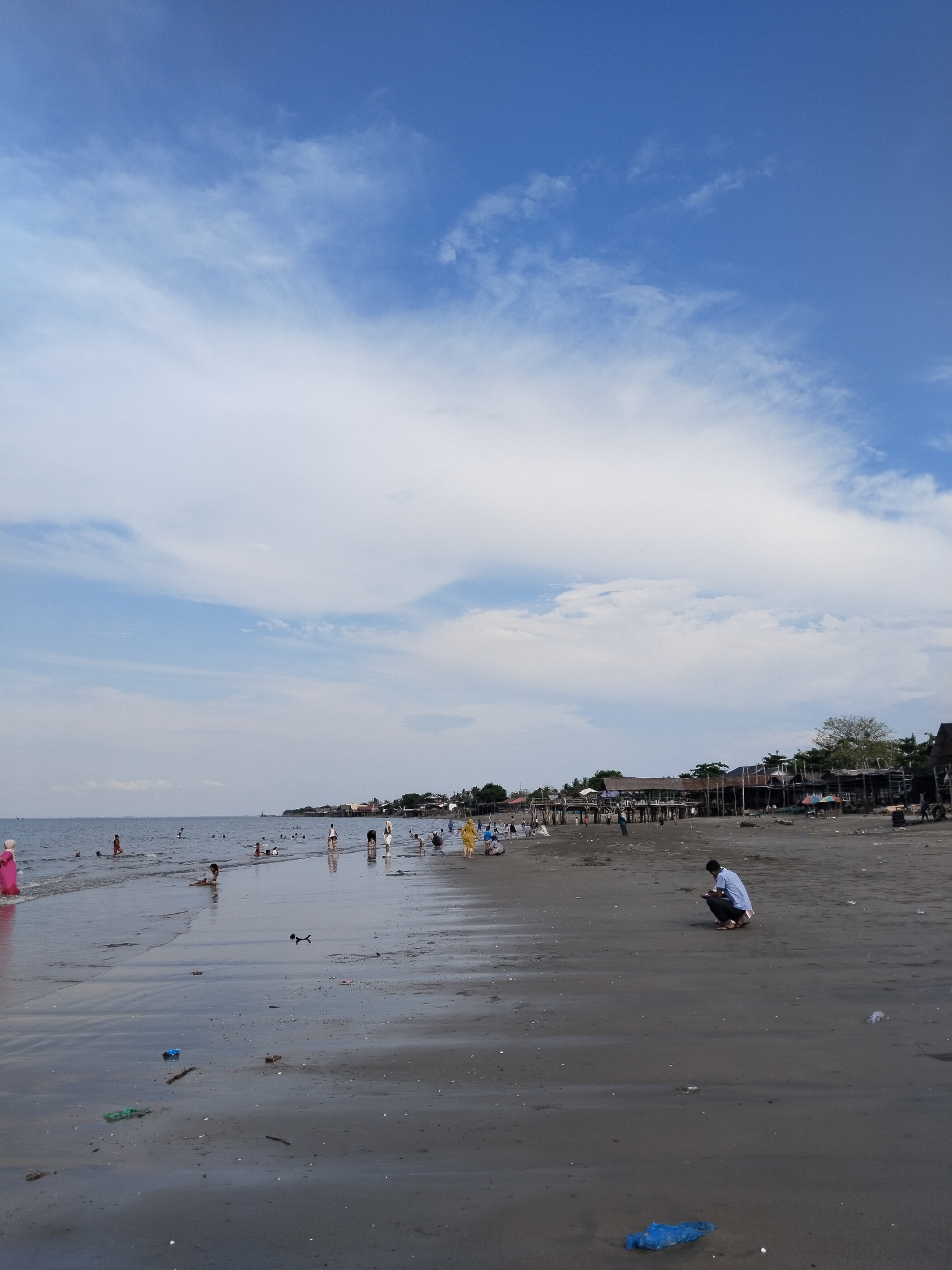 This image depicts a bustling beach scene in Lhokseumawe, Indonesia, under a bright, partly cloudy sky. The horizon divides a vast expanse of calm blue sea from a dynamic sky filled with white, wispy clouds, with patches of clear blue visible above.
In the foreground, a wide sandy beach stretches towards the water, showing reflections of the sky and subtle textures from the receding tide. Numerous people are engaged in various activities along the shoreline. Many individuals, including children and adults, are seen wading, swimming, or playing in the shallow ocean water. Others are walking leisurely along the wet sand. On the right side of the foreground, a person dressed in a light blue shirt and dark pants is prominently featured, crouched down on the sand, appearing to observe or interact with something on the ground.
Along the distant coastline, a series of informal structures, possibly wooden shacks or stilt houses, line the shore, suggesting a local settlement or fishing village. Small boats, one with a visible sail, are dotted on the distant water. The beach itself shows signs of human presence, including some scattered litter like plastic bags. The scene conveys a sense of everyday life and leisure by the sea during what appears to be a pleasant daytime, possibly morning or afternoon, with diffused lighting. No visible text is present in the image.