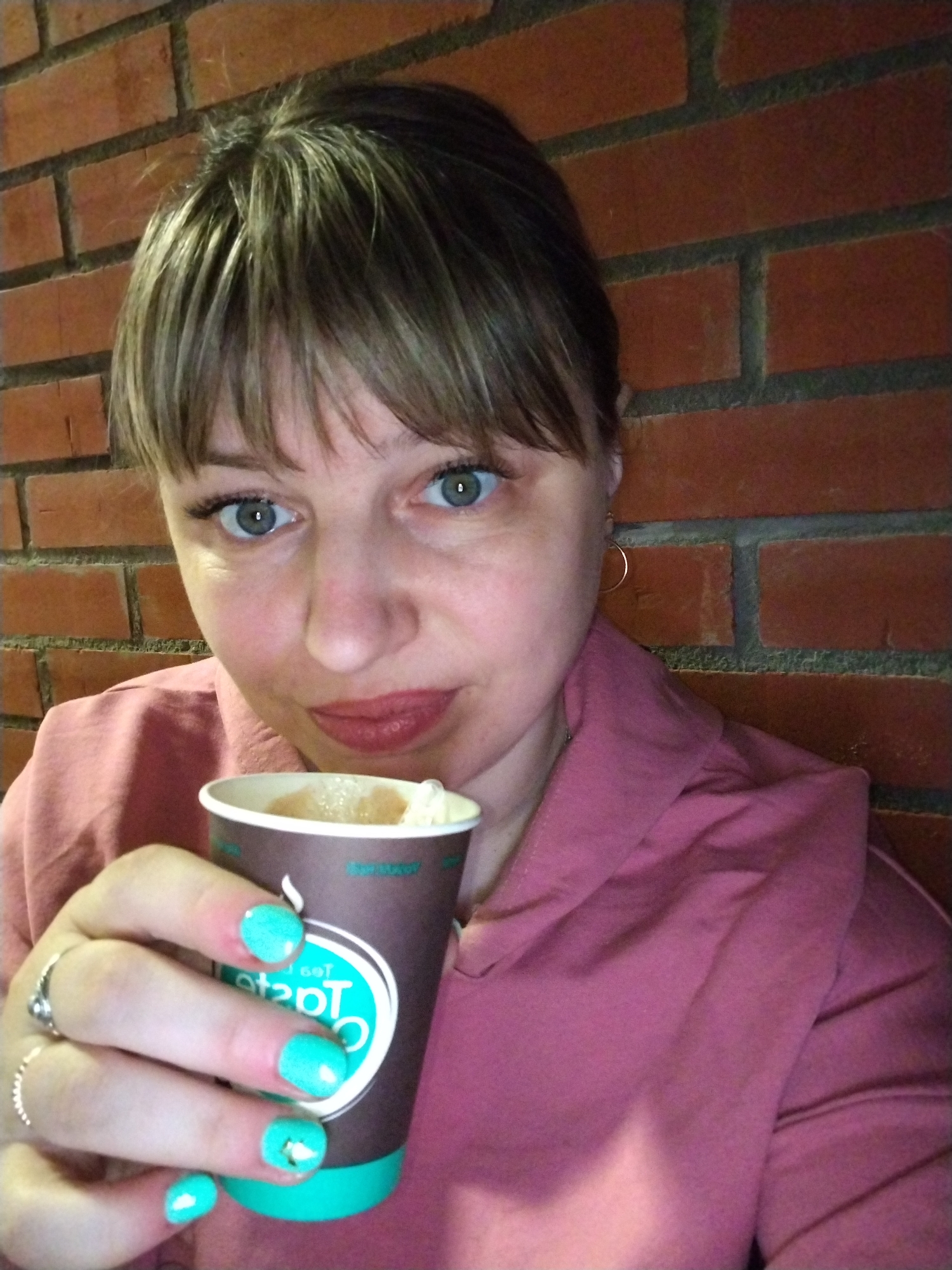 A woman poses for a close-up shot against a red brick wall, likely indoors. She has light brown hair with bangs, prominent blue-green eyes, and is wearing a pink or plum-colored top. Her lips are painted a muted red. She holds a brown disposable cup with her left hand, which contains a light-colored beverage, possibly coffee or tea, topped with foam or whipped cream. Her nails are painted a vibrant turquoise or teal, and she wears two rings on her fingers. A small golden hoop earring is visible on her right ear. The cup features a light blue logo with text, partially visible as "Teatime" or "TeaTaste". Her expression is neutral, looking directly at the camera. The brick wall provides a rustic backdrop. The location context indicates this image was captured in Шебунино, Russia.