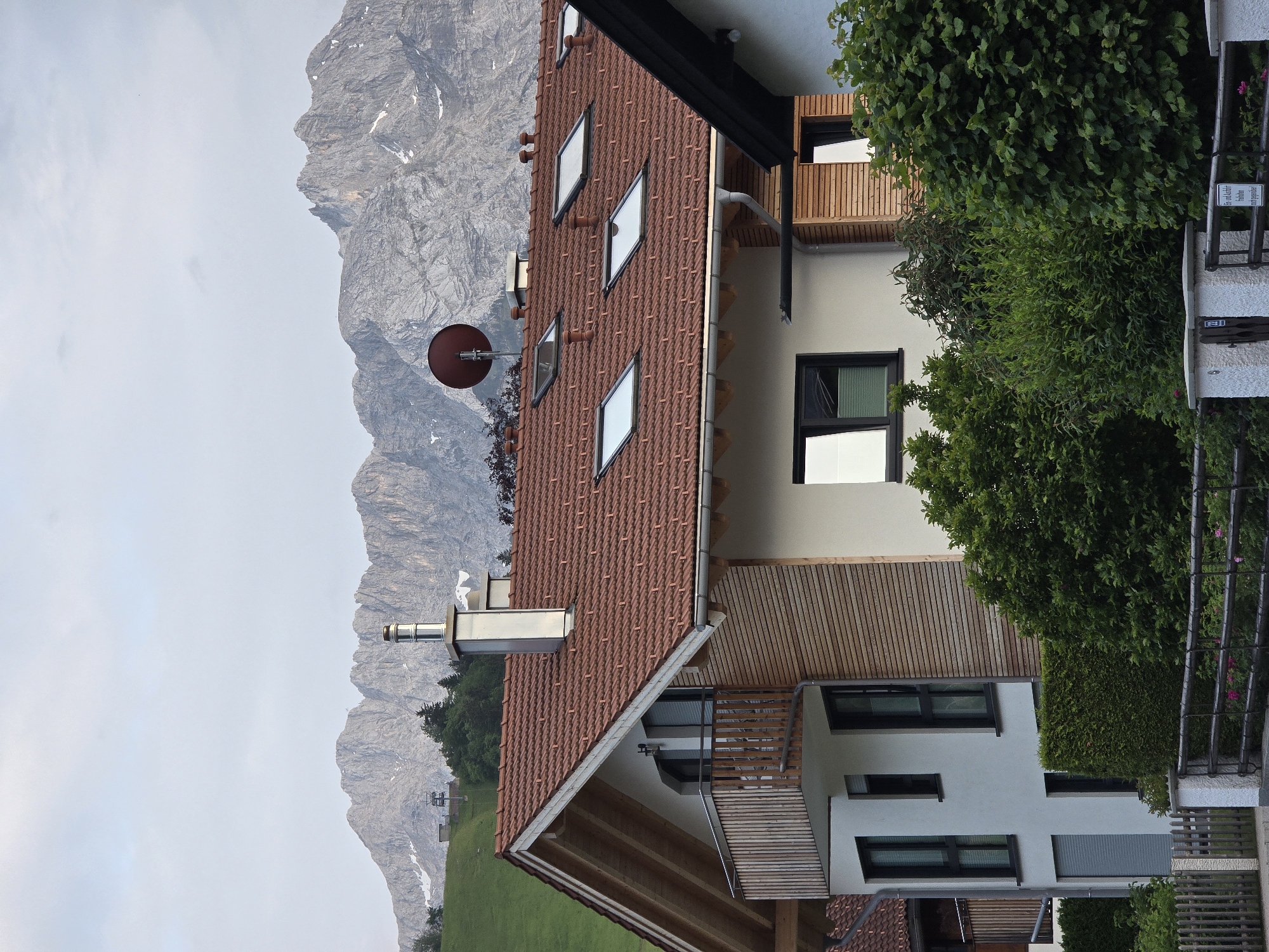The image shows a modern house with a red tile roof in Garmisch-Partenkirchen, Germany. The house features large windows, a balcony, and a wooden facade. In the background, majestic mountains rise under a cloudy sky. A ski lift is visible on the green mountainside to the left, suggesting a ski resort nearby. The scene is tranquil and depicts a scenic mountain village setting. There are no people in the image.