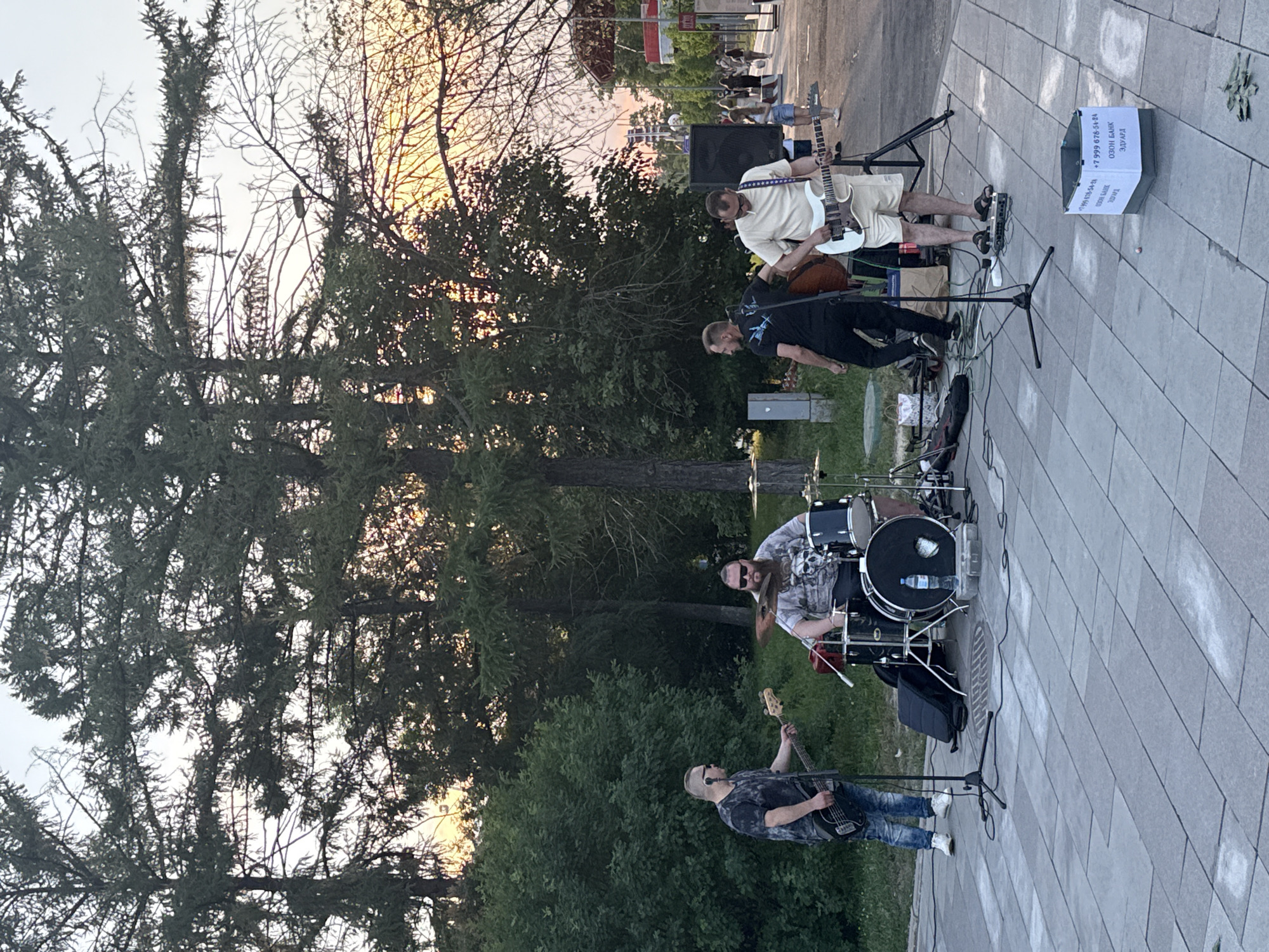 The image depicts an outdoor live music performance in Moscow, Russia. Four men are visible, arranged as a band. On the left, a man with sunglasses and a dark t-shirt plays a black bass guitar. Beside him, a bearded man with sunglasses and a skull-patterned t-shirt sits behind a drum set. To the right of the drummer, a man in a black t-shirt with a white design plays a cello or double bass. Further to the right, a man in a light-colored t-shirt and shorts plays a white electric guitar. The band is set up on a paved walkway, with a grassy area and trees in the background. The sky is a warm sunset hue. In the foreground, a white collection box with text is visible, along with some scattered money. In the background, other pedestrians are present, and a Ferris wheel or similar amusement ride is visible in the distance.