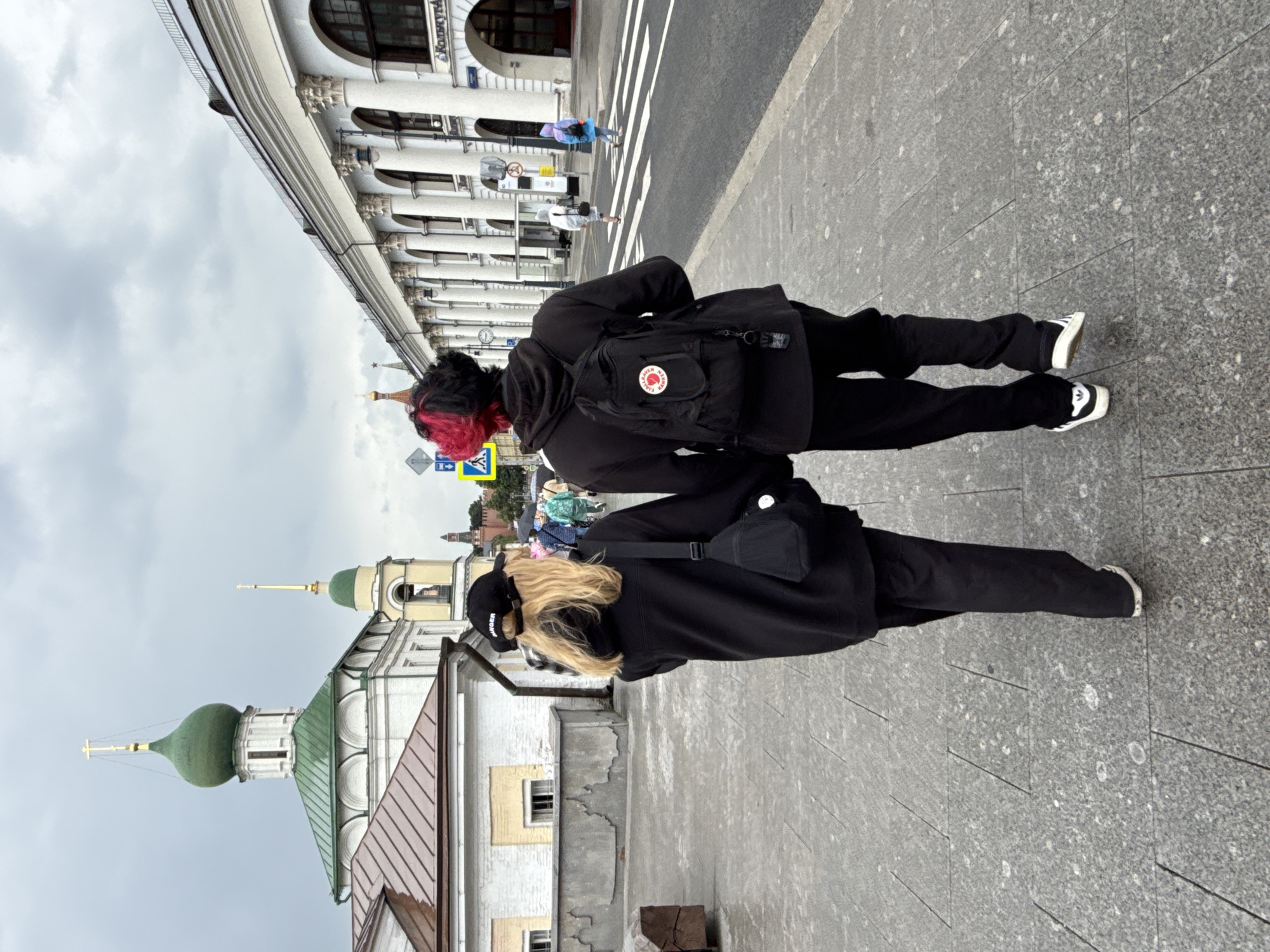 The image shows two people walking away from the camera on a cloudy day in Moscow, Russia. They are walking on a paved sidewalk, and in the background, there is a street with cars and pedestrians. To the left, there is a white building with a green dome and a cross, which appears to be a church. To the right, there is a long, white building with arches, possibly a market or a shopping arcade. The people are dressed in black, and one of them has brightly colored pink and black hair. The overall atmosphere is urban and somewhat somber due to the cloudy weather.