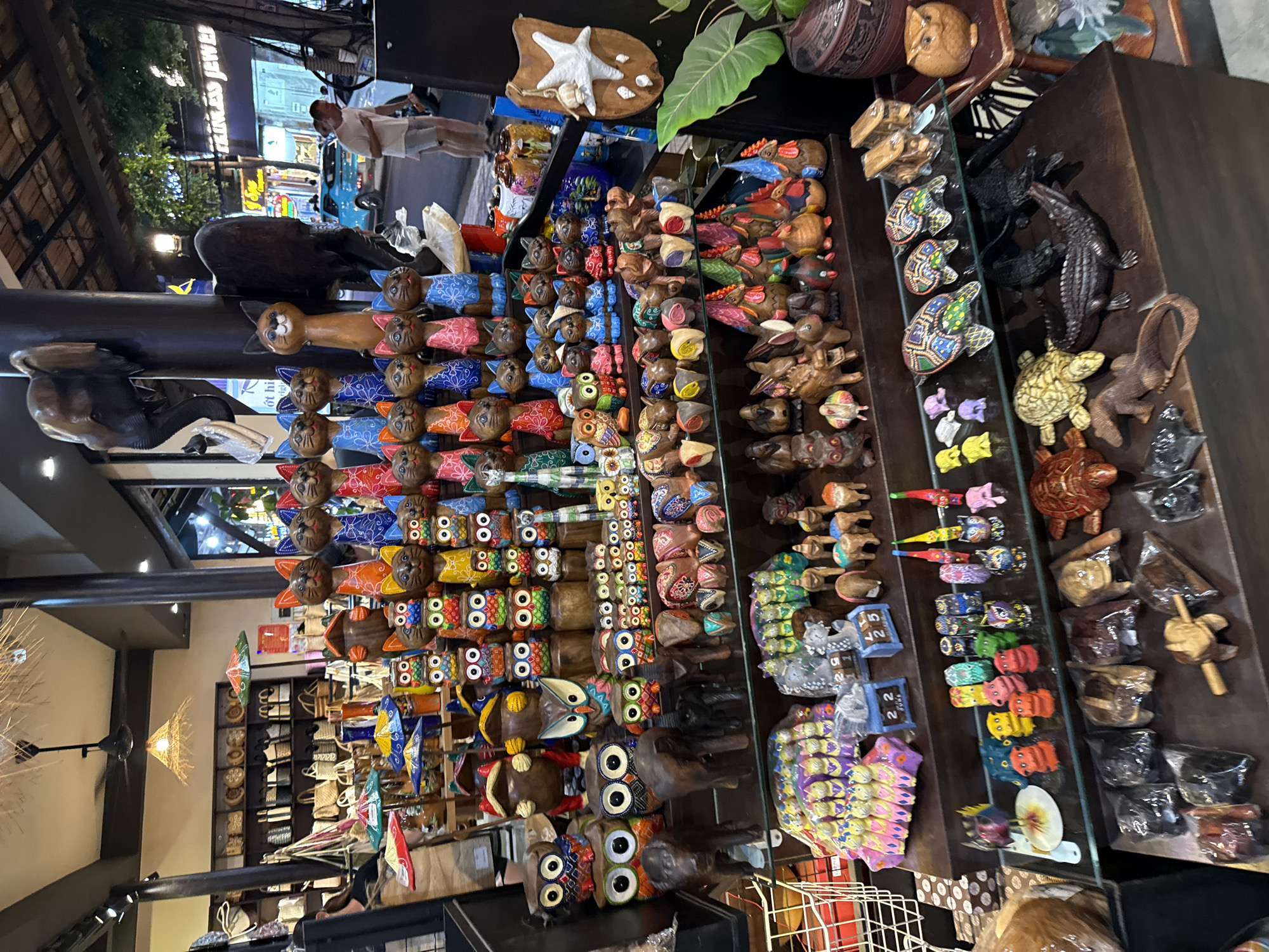 In Nha Trang, Vietnam, a street-side shop displays a variety of handcrafted wooden figurines. The foreground is dominated by a display of dark wooden animal figurines, including crocodiles, lizards, and turtles. Behind this, shelves showcase an assortment of colorful wooden carvings, featuring owls, cats, and roosters, alongside other decorative items. In the background, the shop extends to display woven bags and baskets on shelves. A man in a white t-shirt and shorts is visible walking past the shop entrance on the right. The environment appears to be an outdoor market or commercial street, with a view of other shops and a street in the distance, suggesting a bustling urban setting.