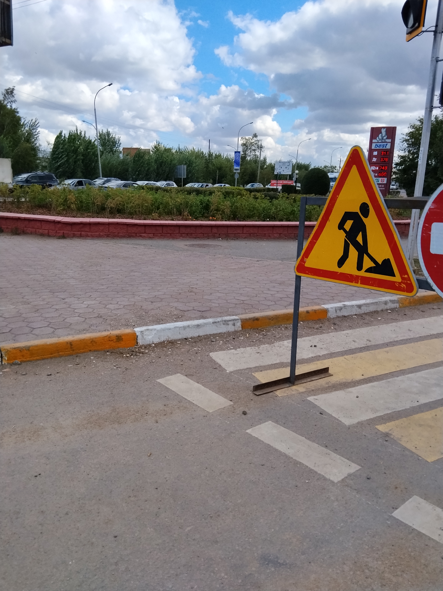 In Kokshetau, Kazakhstan, on a cloudy day, a temporary traffic sign depicting a person shoveling is positioned on a pedestrian crossing. The sign is yellow with a red border and a black silhouette of a worker. Adjacent to it, a gas station sign with prices for various fuels is visible, including "best" branded products. Cars are parked in the background, and a line of trees and bushes borders the road. The pedestrian crossing is marked with white and yellow stripes on asphalt. A curb painted with orange and white stripes is visible to the left of the crossing.