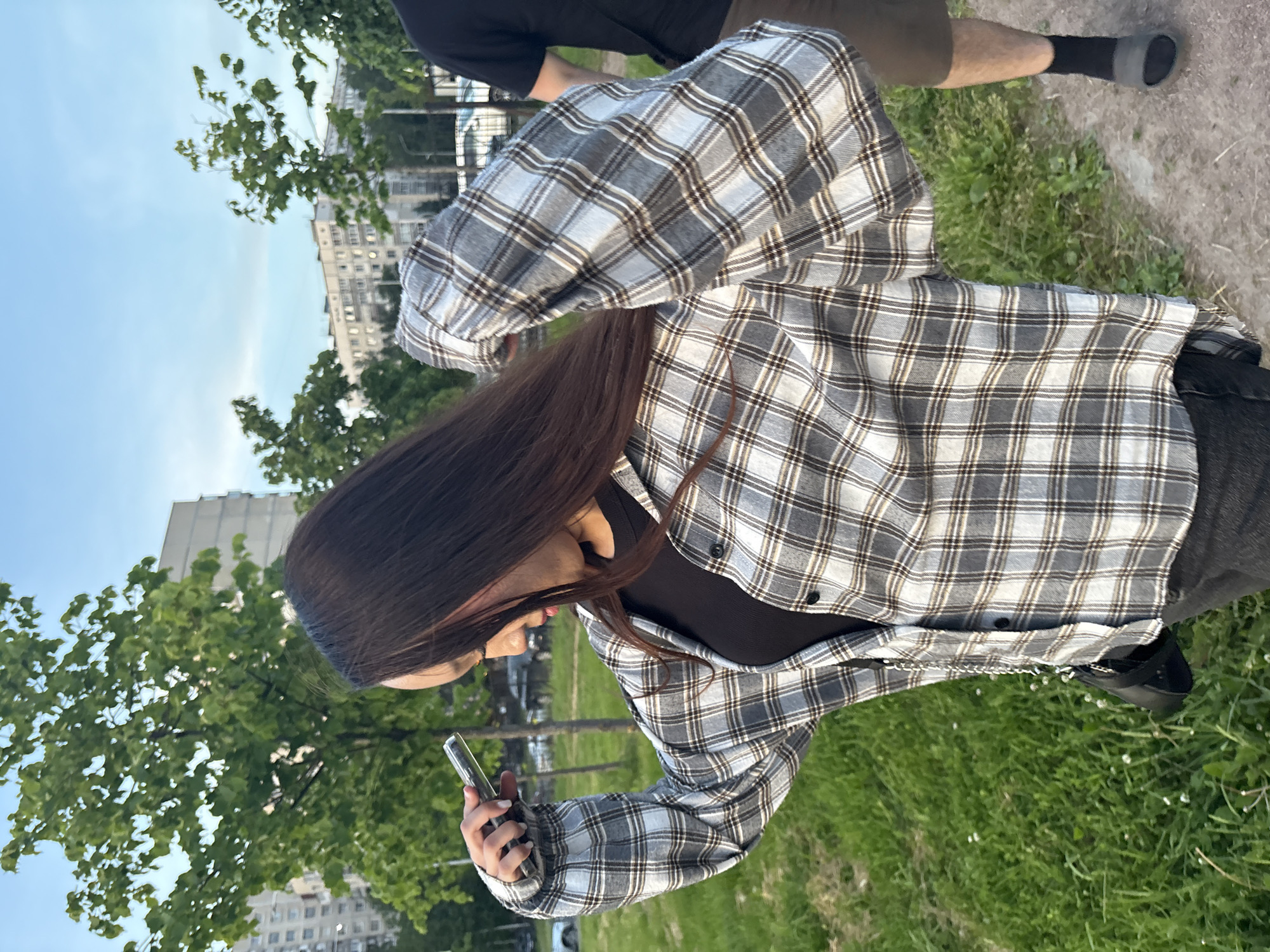 A young woman with long, dark hair stands outdoors in Sinyavino, Russia, in front of buildings and trees. She is wearing a black t-shirt and an open grey and white plaid button-down shirt. Her arms are raised, and she is holding a smartphone in her right hand, seemingly taking a picture or video. A black purse with a chain strap is visible hanging from her left shoulder. The background includes green grass, a paved path, trees with green leaves, and several apartment buildings. The sky is partly cloudy. A person's legs and feet, wearing shorts, dark socks, and grey slides, are visible on the right side of the frame, suggesting they are walking alongside the woman.