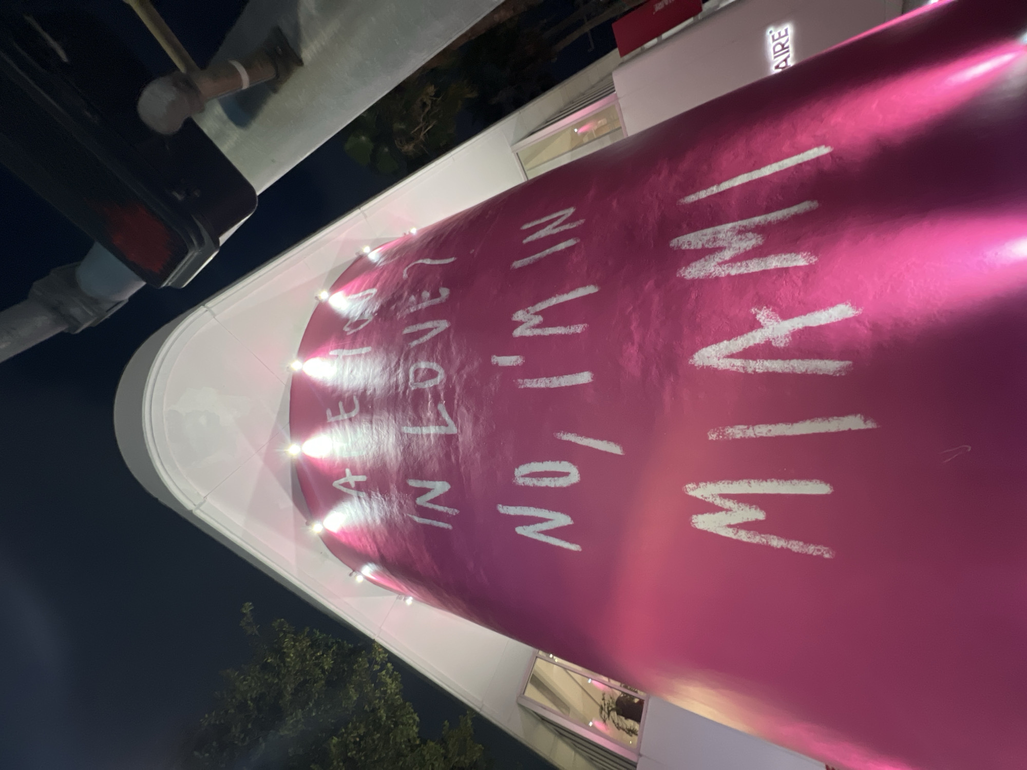 The image depicts a cylindrical pink structure in Miami, USA, bearing the text "ARE YOU IN LOVE? NO, I'M IN MIAMI". The structure is illuminated by lights at the top, and a building with white walls is visible in the background. Some trees are also noticeable. In the upper left corner, a portion of a street light is seen. The image was taken at night.