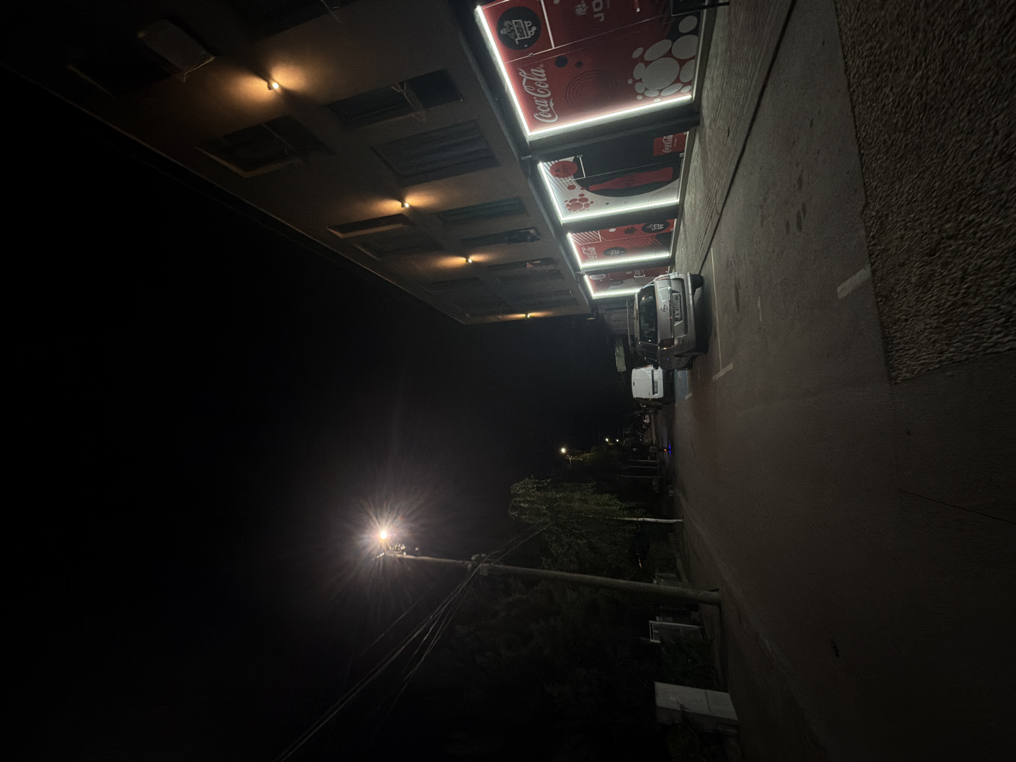 The image depicts a nighttime street scene in Obzor, Bulgaria. A modern, two-story building with a neutral-toned facade dominates the right side of the frame, its upper floors illuminated by several external wall lights. The ground floor features large, illuminated storefronts adorned with Coca-Cola branding and circular graphics. A silver sedan is parked in front of the building, its license plate visible. A white van is parked further down the street on the right.
The street itself is paved and shows signs of wear, with white lines marking parking spaces. A tall streetlamp with a bright, starburst-like light source is positioned on the left side of the road, casting light onto the street and overhead power lines. Adjacent to the streetlamp is a dense line of trees, some of which appear to be weeping willows. Further down the street, another streetlamp is visible, and a person is seen riding a scooter or electric unicycle. The overall environment suggests a quiet commercial or residential area at night.
