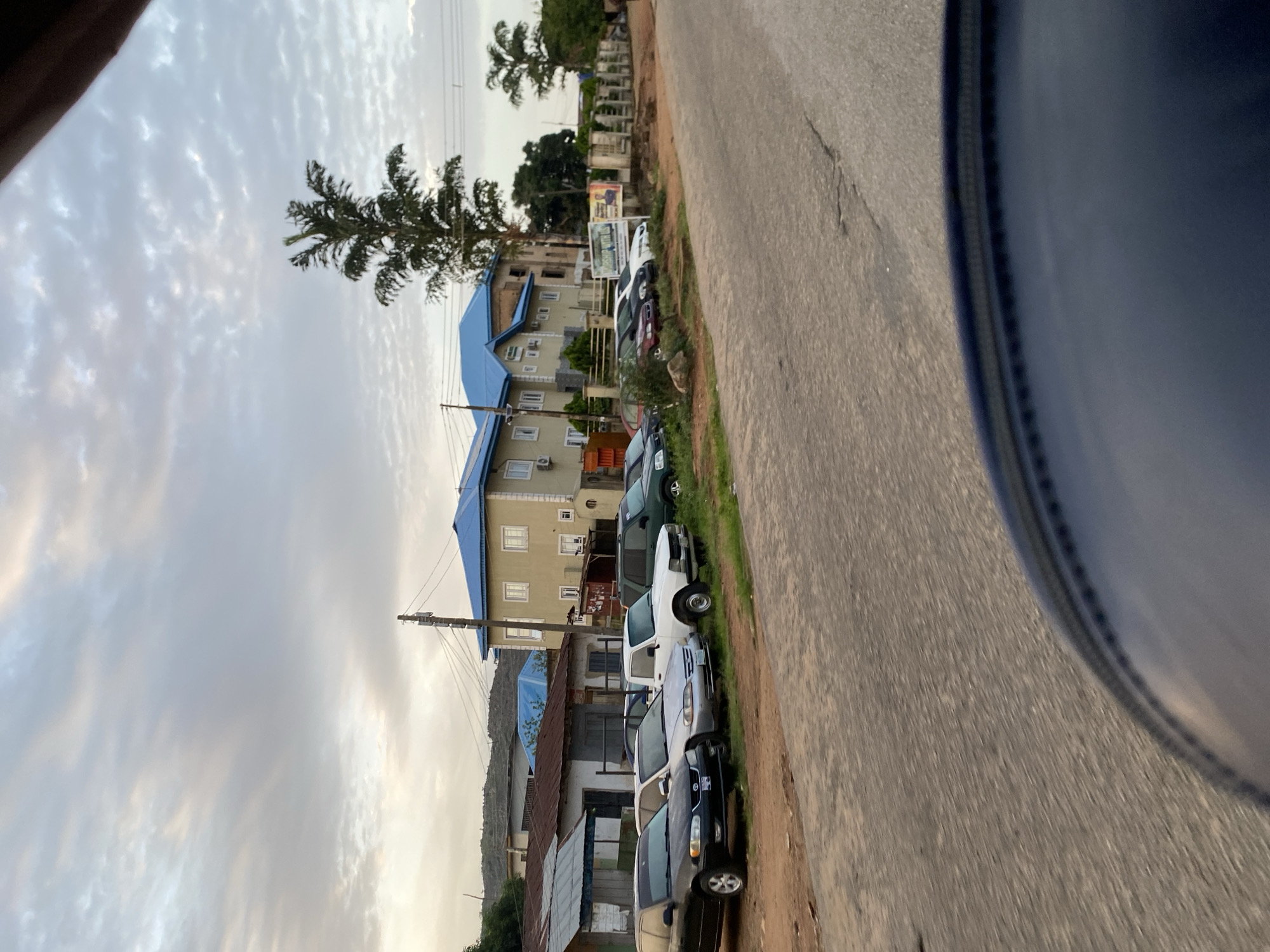 The image depicts a street scene in Jos, Nigeria, likely during a late afternoon or early evening given the soft light and overcast sky. A row of parked cars lines the side of a paved road, with several buildings visible behind them. The architecture of the buildings varies, ranging from what appears to be older, single-story structures with corrugated metal roofs to a more modern, multi-story building with a bright blue roof. A tall, slender tree with sparse foliage stands prominently near the multi-story building. The road surface is asphalt, showing some wear and tear. The foreground is partially obscured by what seems to be the edge of a dark-colored object, possibly part of a vehicle from which the photo was taken. The overall impression is of a typical urban street in a developing area, with a mix of old and new construction and a functional, albeit somewhat unkempt, environment. No people are clearly visible in the image.