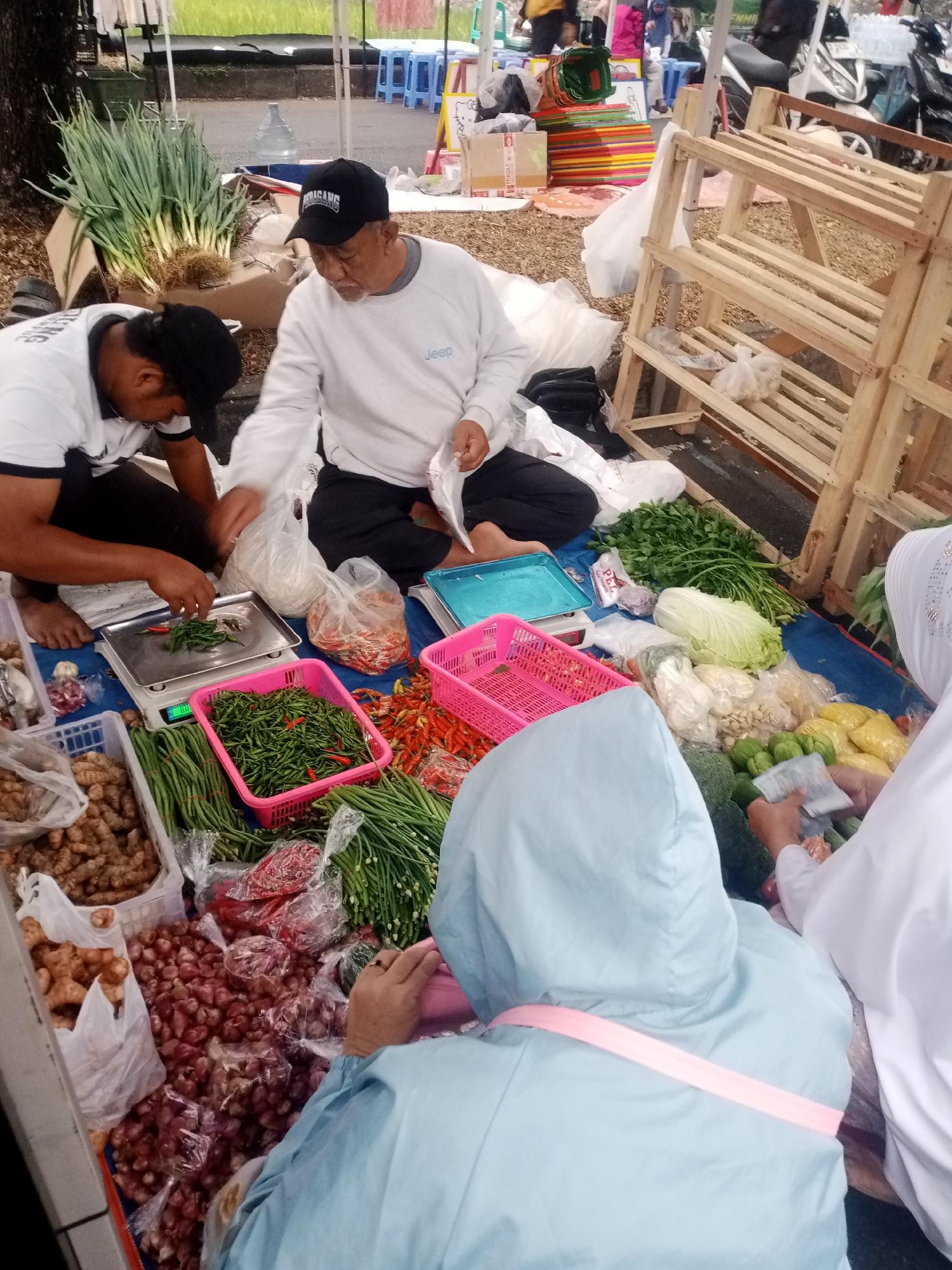 This image depicts an outdoor market scene in Tasikmalaya, Indonesia, likely a wet market or a roadside stall, with a variety of fresh produce displayed on the ground and on wooden crates. The focus is on two individuals, presumably vendors or customers, engaged in a transaction involving weighing green chilies on a digital scale.
In the foreground, a man wearing a white long-sleeved shirt and a black baseball cap is seated and appears to be assisting with the weighing of chilies. To his left, another person, also wearing a white shirt with a black collar and a black baseball cap, is also seated and focused on the produce. A variety of vegetables are visible, including large bunches of green onions, long beans, red onions in plastic bags, ginger, cabbage, and various types of chilies in red and green colors, some in pink plastic baskets.
In the mid-ground, a wooden shelving unit with multiple tiers stands empty, suggesting it might be used for display or storage. Further in the background, other market activities are hinted at, with blurred figures and stalls, along with a parked motorcycle. The ground is covered with a blue tarp, which serves as the base for the displayed produce. The overall lighting suggests it is daytime, possibly overcast or in a shaded area of the market. The presence of customers wearing headscarves indicates a likely cultural context for Indonesia.