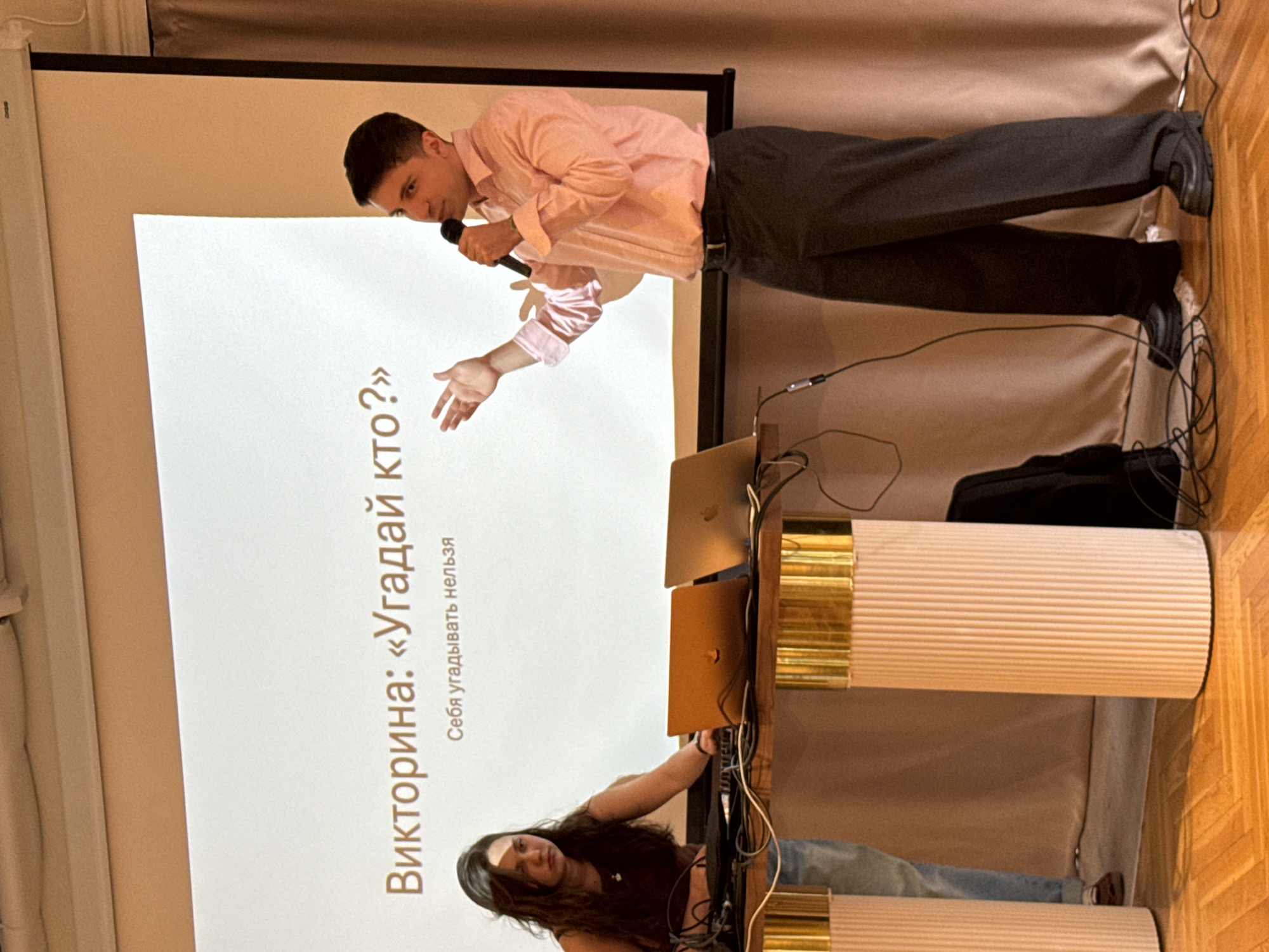 The image depicts a presentation or event at an indoor venue in Khimki, Russia. A man in a light pink collared shirt and dark trousers stands on the right, holding a microphone and gesturing with his left hand. He appears to be speaking to an audience, though they are not visible. A projection screen behind him displays text in Russian: "Викторина: «Угадай кто?»" which translates to "Quiz: 'Guess Who?'" Below this, smaller text reads "Себя угадывать нельзя" meaning "You cannot guess yourself." On the left, a woman with long dark hair, wearing a dark sleeveless top and blue jeans, stands behind a table with two laptops and other equipment, looking down. The table is supported by two cylindrical pedestals with decorative ribbed texture and gold-colored tops. The room has wooden flooring and a light-colored wall or curtain in the background. The overall atmosphere suggests a professional or educational event.