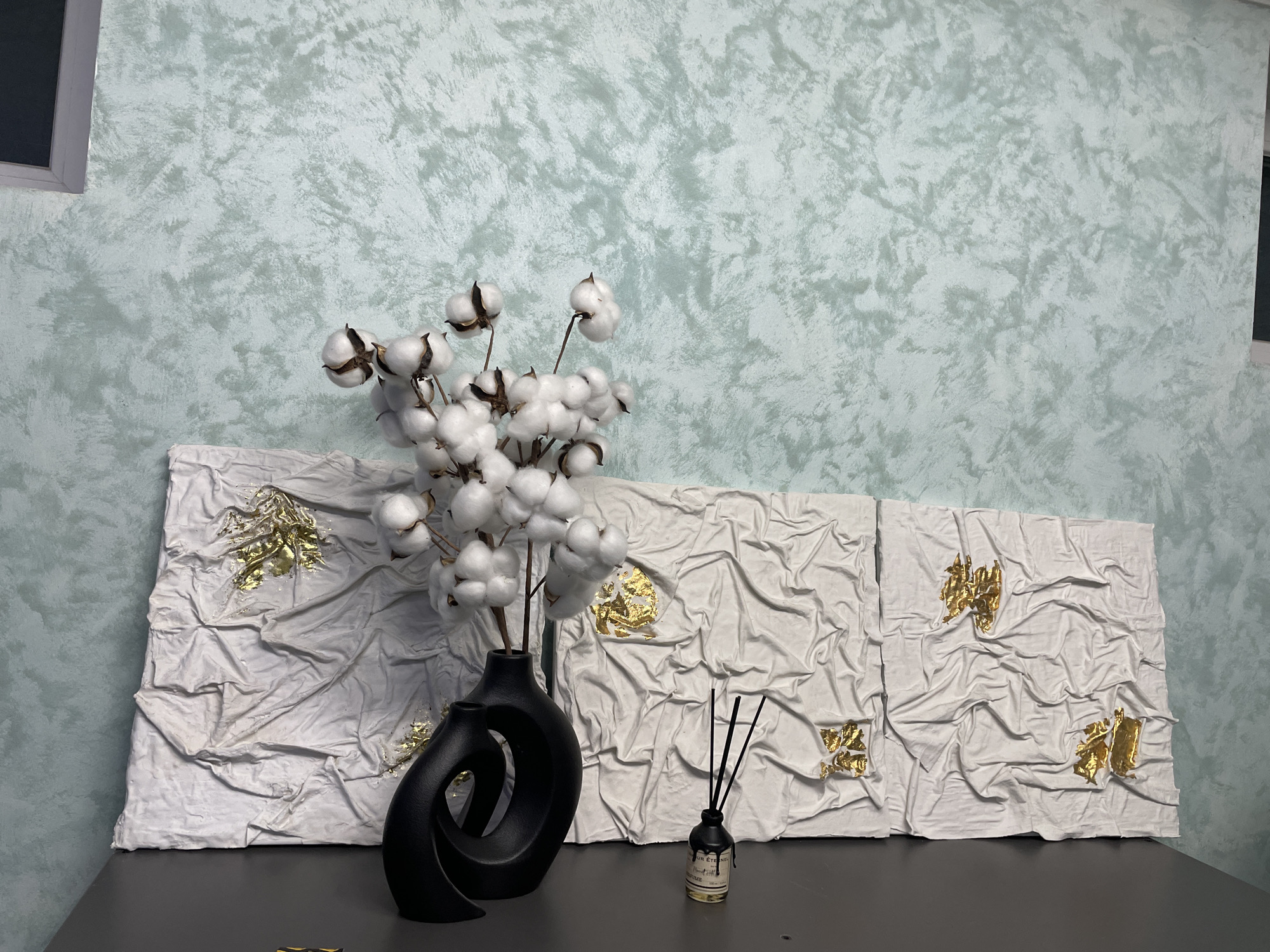 The image depicts an indoor scene with a focus on decorative objects placed on a dark gray table. In the foreground, a striking black vase with a unique, stylized design holds several cotton stems with fluffy white cotton bolls. To the right of the vase sits a small, dark-labeled bottle with black reed diffuser sticks protruding from it.
Behind these objects, a triptych artwork is displayed. Each panel of the artwork features a textured white surface with gold leaf embellishments scattered across it. The texture of the white surface appears to mimic the look of crumpled fabric.
The background consists of a wall with a mottled, textured surface in shades of light blue and gray. A portion of a window frame is visible on the left side of the image.
The image was taken in Лесной Пейзаж Кп (шарапово), Russia.
