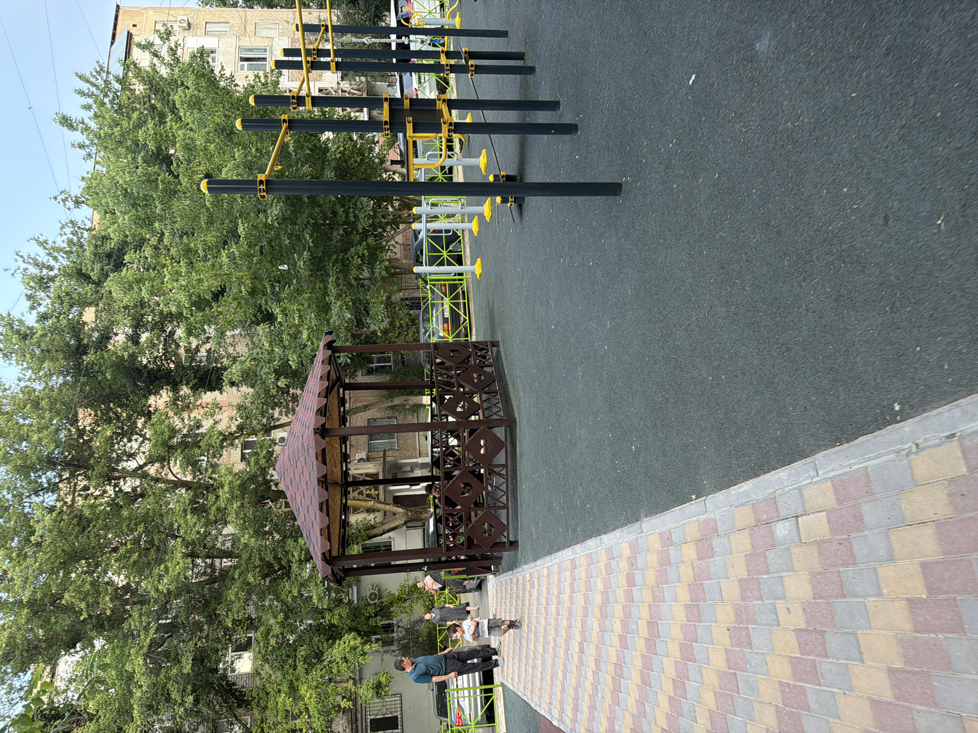 The image depicts an outdoor recreational area in Shymkent, Kazakhstan, featuring a modern playground with a gazebo and exercise equipment. In the foreground, a paved walkway with a checkerboard pattern of beige, pink, and grey tiles leads towards a dark brown, octagonal gazebo. The gazebo has a red, shingled roof and a decorative metal fence. To the right of the gazebo, a black rubberized surface forms the playground ground. A set of black metal workout bars with yellow accents is prominently displayed on the right side of the image, suggesting a fitness zone.
In the background, several people are visible. A man in a dark blue shirt and black pants stands near the walkway, facing a young child in a light-colored t-shirt and grey shorts who is walking away from the camera. Behind them, another woman in a dark dress and a headscarf is also present. Further in the background, apartment buildings with balconies and windows are visible, interspersed with lush green trees that provide shade. Cars are parked along the street in front of the buildings, some with visible license plates. The sky is a pale blue, indicating daytime. The overall scene conveys a sense of community activity and public space utilization.