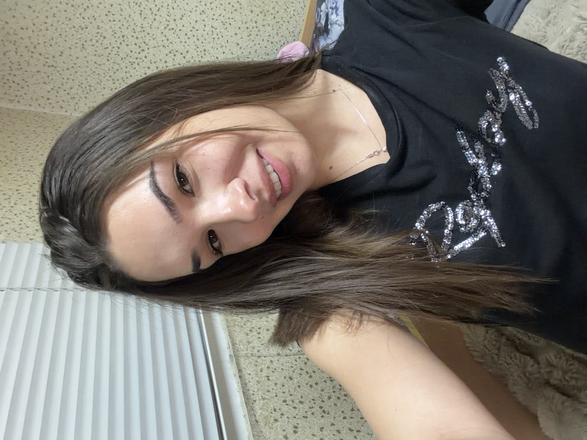 A woman is taking a selfie. She has long brown hair and is wearing a black shirt with silver detailing. The background is a beige wall with a window to the left. Based on the images provided, I cannot determine the location in Sofia, Bulgaria.
