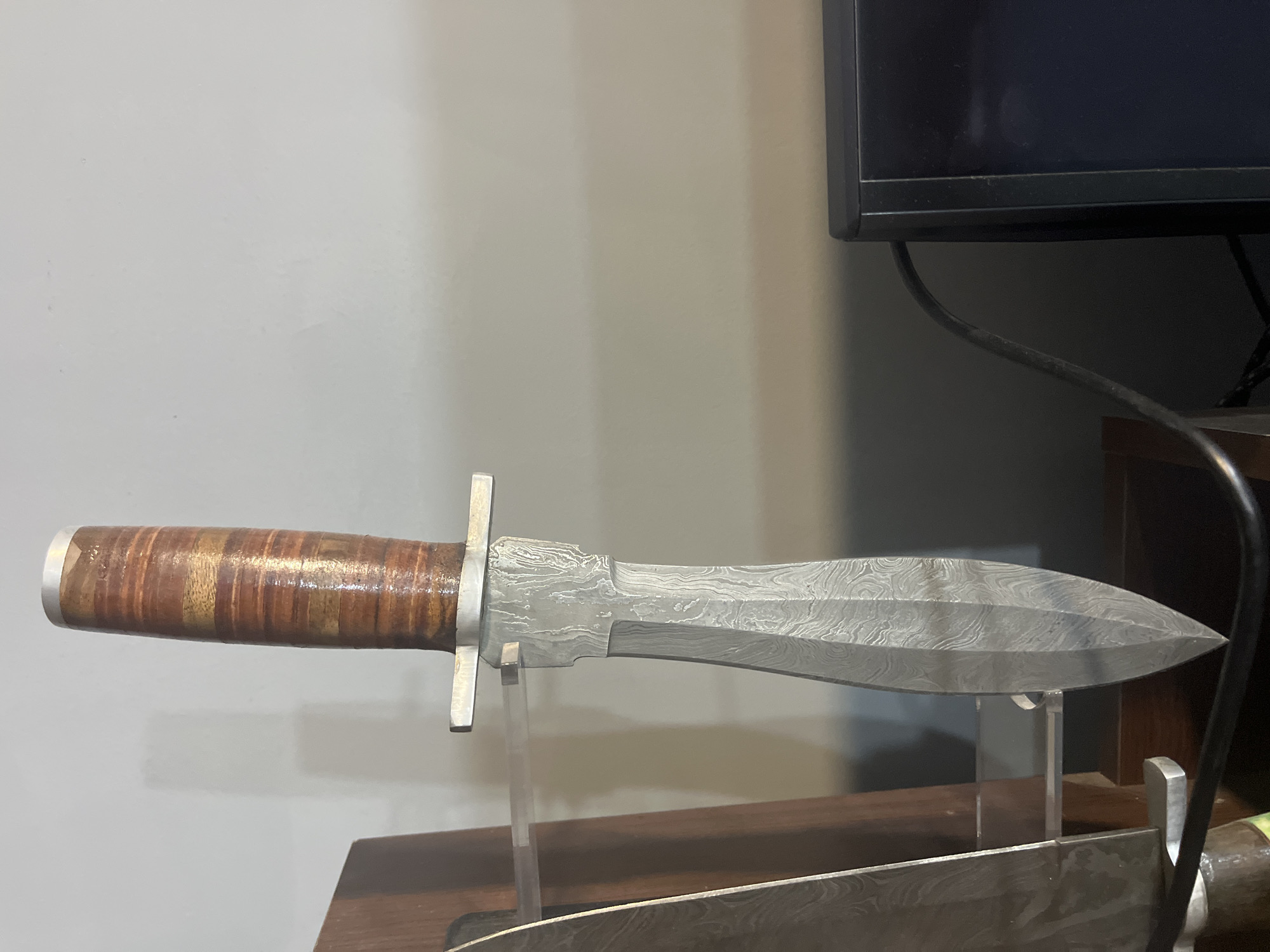 A knife with a patterned steel blade and a layered brown leather handle is displayed horizontally. The knife is supported by two clear acrylic stands, which are placed on a wooden surface. In the background, a portion of a dark computer monitor is visible, along with a black cable. The wall behind the monitor and the knife is a light, neutral color. The image is an indoor shot. The location is not specified in the city of Montana, Bulgaria.