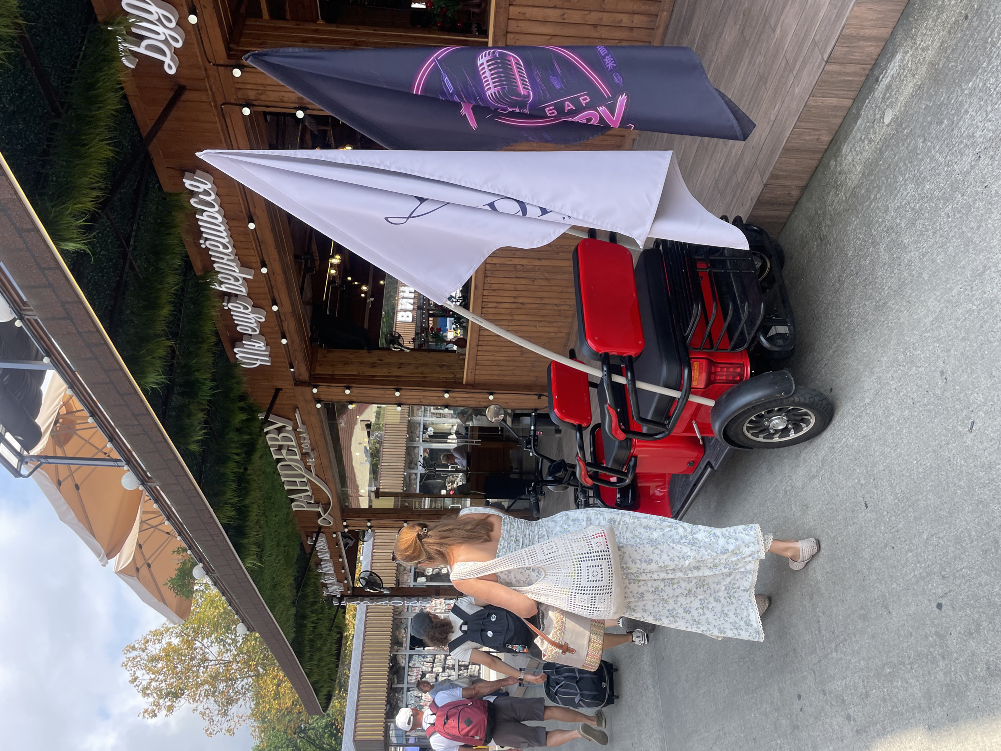 In Sochi, Russia, on a bright, overcast day, a red electric scooter with two seats and a basket sits parked on a paved walkway in front of a wooden building with a green, grass-like facade and beige awnings. Two flags, one white with a dark inscription and one black with a neon pink microphone graphic and text, are attached to the scooter. Behind the scooter, several people walk away from the camera. A woman in a long, floral print dress and a woven tote bag is in the foreground, facing away from the camera. Two other individuals with backpacks are walking ahead of her, and a man with a red backpack is further ahead. The building appears to be a cafe or restaurant, with large glass windows and illuminated signage in Russian.
