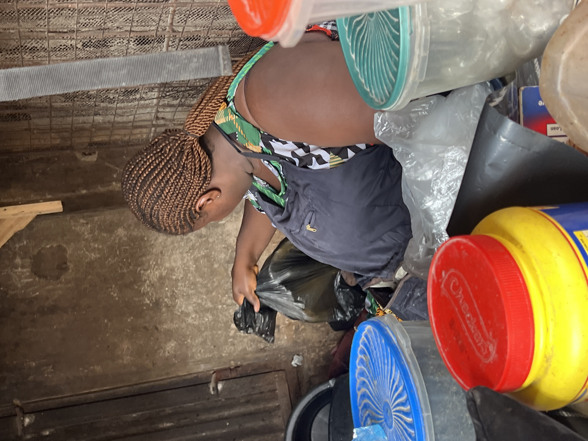 In Nkpor, Nigeria, a woman with braided hair is seen from behind, wearing a dark apron over a patterned top. She is holding a black plastic bag filled with contents. The environment appears to be a store or a storage area, with various containers visible, including a yellow and red container with a "Checkers" logo on its lid, and clear plastic containers with blue and teal lids. The background shows a textured, grimy wall and a wire mesh structure. The lighting suggests an indoor or shaded outdoor setting.