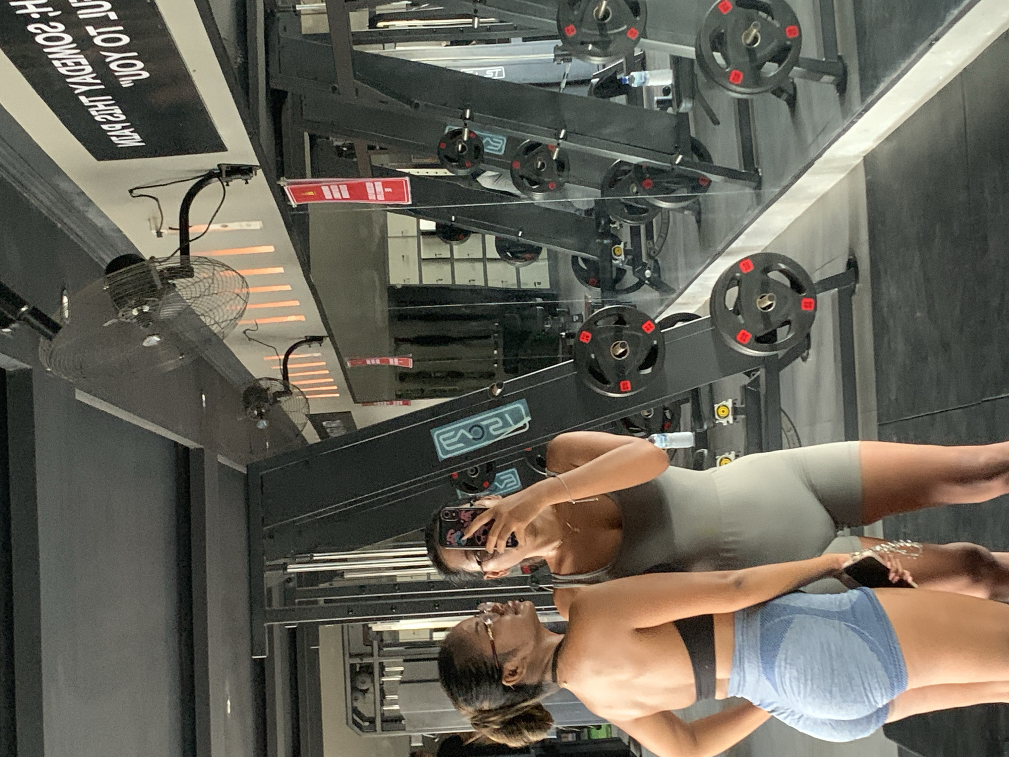 Two women stand in front of a mirror at a gym in Sukawati, Indonesia. One woman, wearing blue shorts and a black sports bra, has her back to the mirror and is slightly turned towards the camera. The other woman, wearing a grey sports outfit, holds a mobile phone up to take a photo of their reflection in the mirror. Gym equipment with black weights is visible in the reflection. A fan is mounted on the ceiling.
