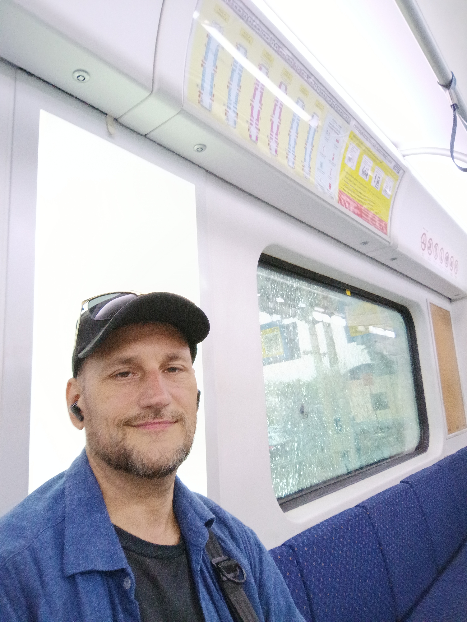 A man wearing a blue collared shirt and a black baseball cap with sunglasses perched on top is seated inside a train. He has a beard and is wearing earbuds. The train's interior is visible, with blue patterned seats and a window showing a blurry, possibly rain-streaked, reflection of the outside. Safety and route information posters are displayed above the window. The man appears to be taking a selfie. The location is the KTM Komuter train service in Klang City, Malaysia.