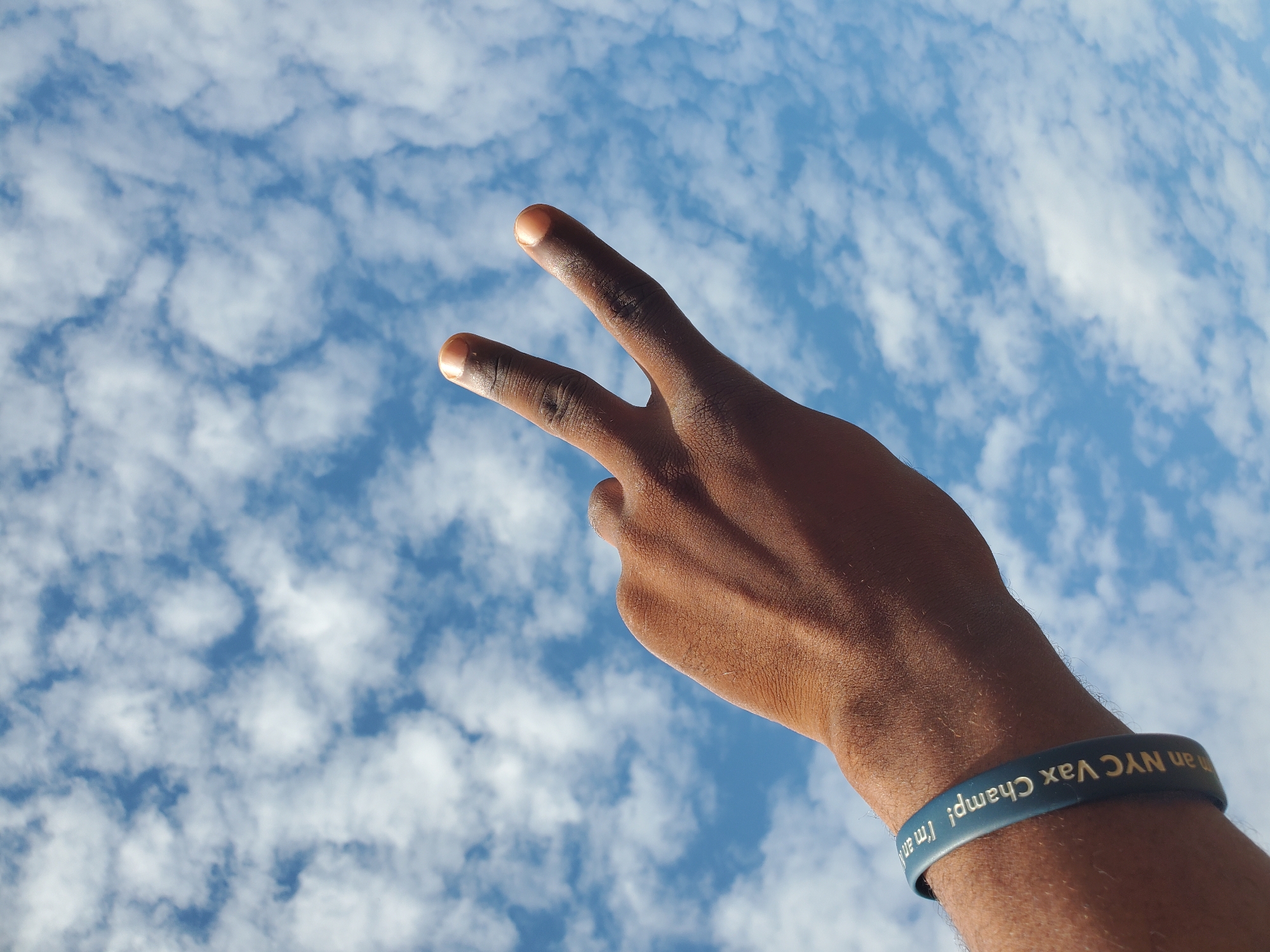 The image depicts a left forearm and hand with dark brown skin, extended upwards against a bright blue sky filled with scattered white cumulus clouds. The index and middle fingers are raised, forming a 'V' shape, while the other fingers are folded. A dark blue silicone wristband is worn on the forearm, featuring gold lettering that reads "I'm an NYC Vax Champ!". Fine hair is visible on the skin of the arm and hand, and the fingernails are clean. The scene is located in the city of Winters, United States of America.