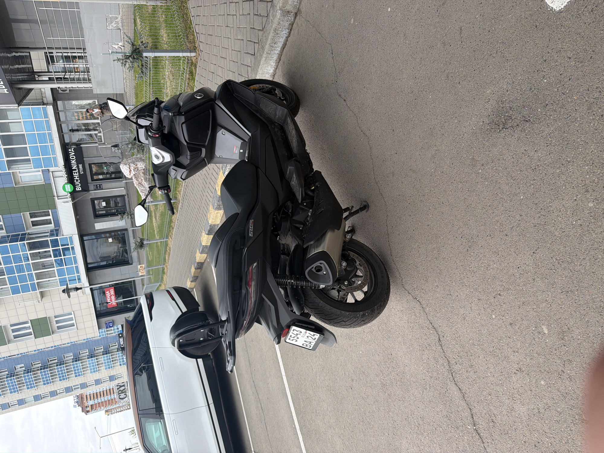 A black scooter is parked on an asphalt surface next to a white car. The license plate of the scooter reads "5543 BA 24," indicating registration in the Krasnoyarsk Krai region of Russia. The surroundings include a building with the sign "BUCHELNIKOVA STORE" and blue-tinted windows, suggesting a commercial or residential area in Krasnoyarsk, Russia. The weather appears to be clear and sunny. There are no visible people actively interacting with the scooter or car. The scooter is angled towards the right side of the frame and positioned on a parking line.