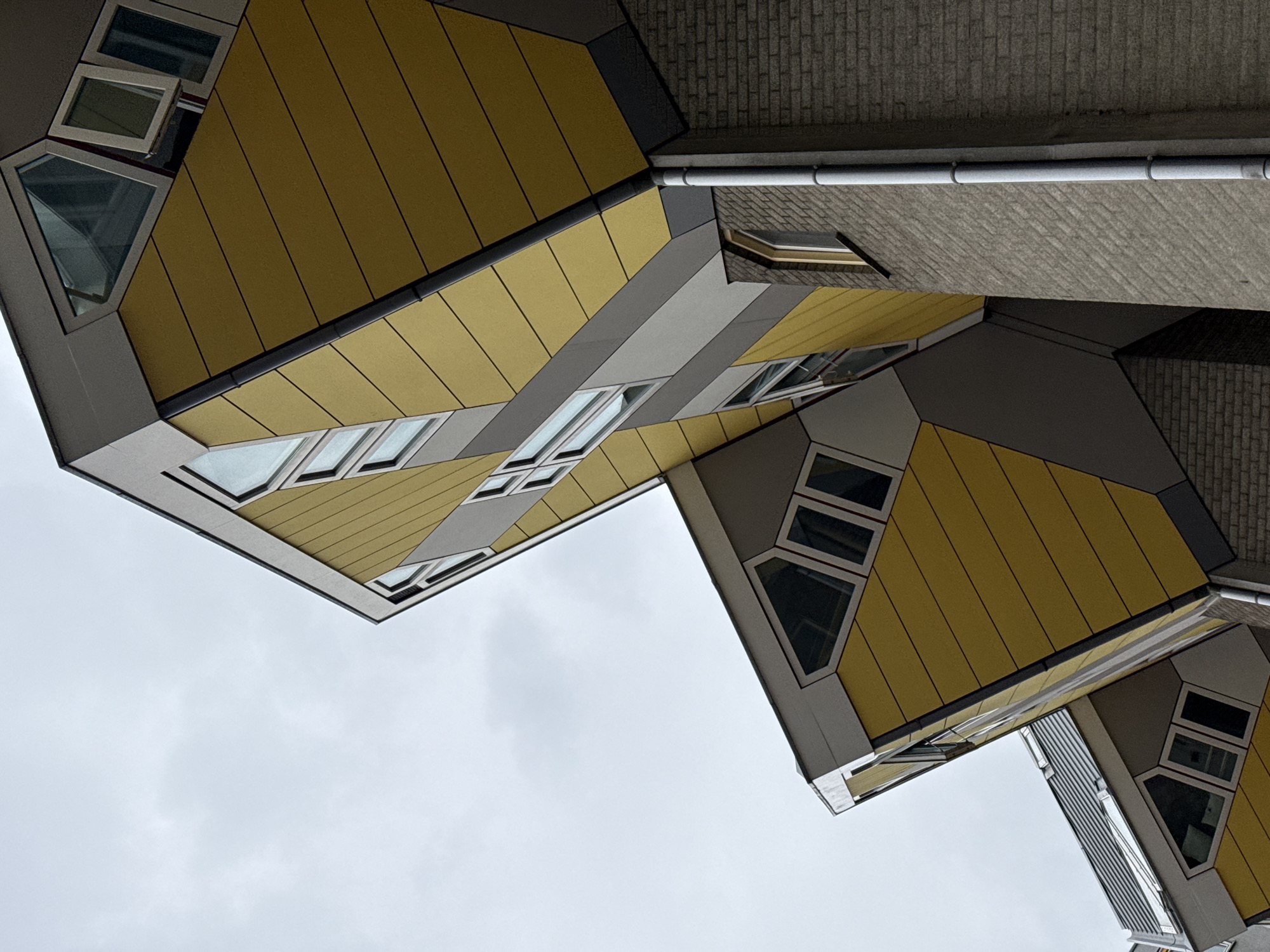 The image captures a low-angle, upward view of the iconic Cube Houses (Kubuswoningen) in Rotterdam, Netherlands. The architecture is characterized by tilted yellow cubic structures with triangular windows, juxtaposed against a gray, overcast sky. The buildings appear to be made of materials like metal siding and brick. A drainpipe runs vertically along a brick wall to the right of the frame. The scene is devoid of people, emphasizing the unique architectural design. The lighting suggests it is daytime, but the cloudy weather creates a diffused and somewhat muted atmosphere.