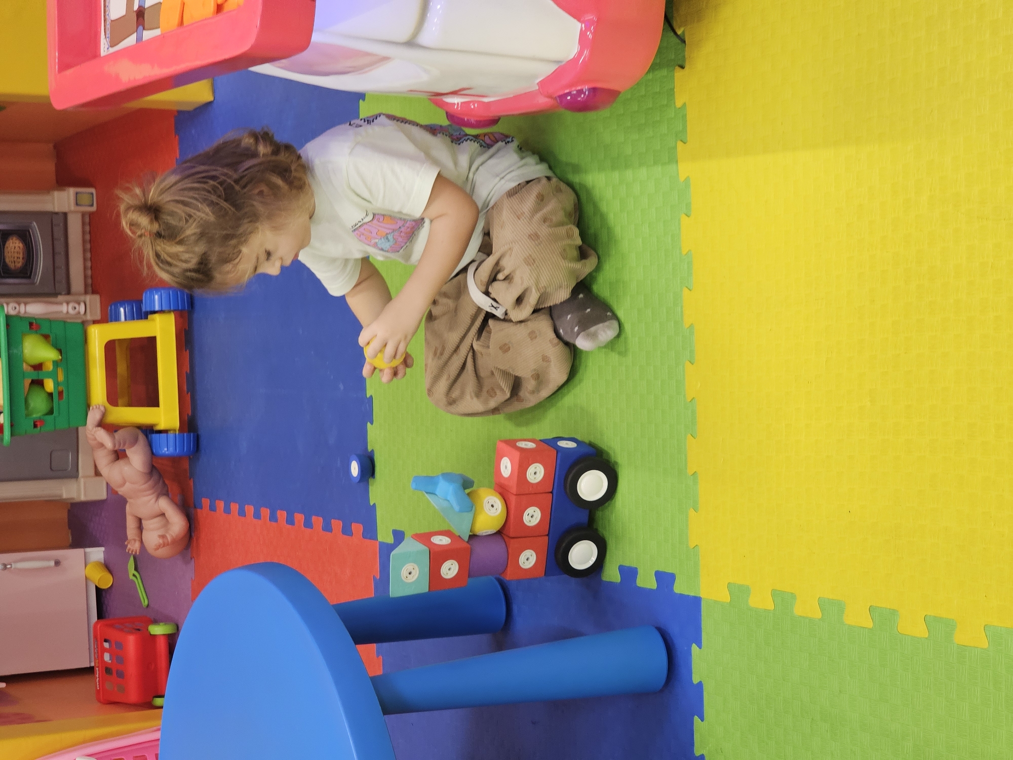 A toddler sits on colorful foam floor tiles in a playroom. The tiles are predominantly green, yellow, blue, and red. The child wears a light-colored t-shirt, brown corduroy pants, and one grey sock. The child holds a yellow ball in their hand. Around the child are various toys, including building blocks, a blue table, a play kitchen set, and a red shopping cart. A baby doll lies near the kitchen set. The location is Khimki, Russia, based on the provided information.