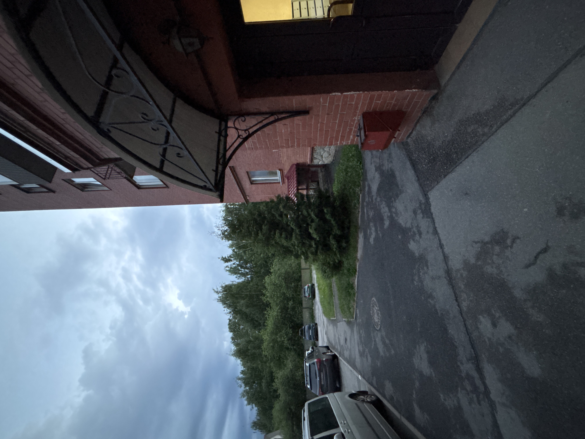 The image depicts an outdoor scene in a residential area of Saint Petersburg, Russia, under overcast skies, suggesting either dusk or a heavily cloudy daytime condition.
In the right foreground and midground, a multi-story building constructed of red-brown brick is prominently featured. It has white-framed windows visible on its upper floors and a lower section. An arched, dark-colored metal canopy with decorative wrought-iron scrollwork extends from the facade, covering an entrance. Below this canopy, a red rectangular metal box is mounted on the brick wall. The entrance itself has a dark-colored door, and an interior light source casts a yellow-orange glow, illuminating what appears to be an office or utility space with visible white, multi-drawer cabinets.
The foreground and midground consist of an asphalt paved area, exhibiting numerous dark, wet-looking patches consistent with recent rain or damp conditions. A circular metal manhole cover is embedded in the asphalt in the midground.
Several vehicles are parked on the asphalt. On the left, a light-colored van or minivan with roof rails is partially visible. Further into the midground, a dark-colored SUV, also with roof rails, is parked facing away from the viewer, displaying a Russian license plate. Another dark-colored SUV is parked behind it. To the right, partially obscured by a tree, a dark-colored car is parked.
The vegetation includes a prominent, dense coniferous tree (likely a spruce or fir) situated on a patch of green grass in the midground, between the asphalt and the background. The background is dominated by a thick line of various green deciduous trees, forming a natural barrier or greenbelt. Additional patches of green grass are visible along the edges of the asphalt.
To the left of the coniferous tree and in the midground, another red-brown brick building is partially visible, featuring white-framed windows and an attached small structure with a red, sloping roof.
The sky is entirely covered with dense, grey clouds, with some lighter grey patches indicating diffused light. No people are present, and no actions or interactions are depicted within the scene. The architectural style, vehicle types, and the discernible Russian license plate format consistently place the location within Saint Petersburg, Russia.