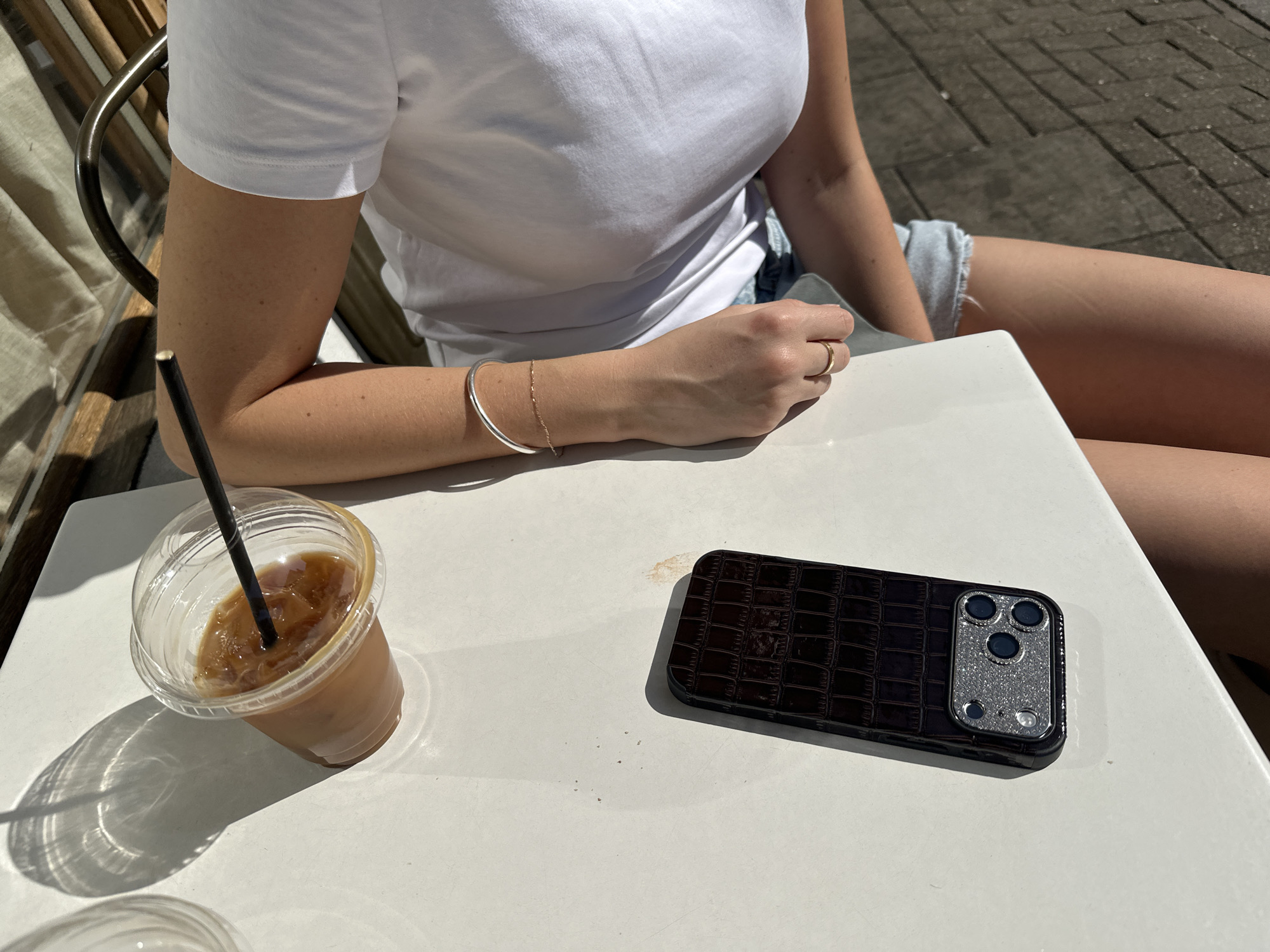 A woman with tanned skin, wearing a white T-shirt and denim shorts, sits at a white table. A plastic cup containing a brown liquid with ice and a black straw sits on the table next to a black phone case with a mock-croc design and a glittery camera surround. The setting is an outdoor café or similar venue with a wooden bench visible in the background and a paved walkway visible in the upper right. The location is not identifiable as London, United Kingdom.