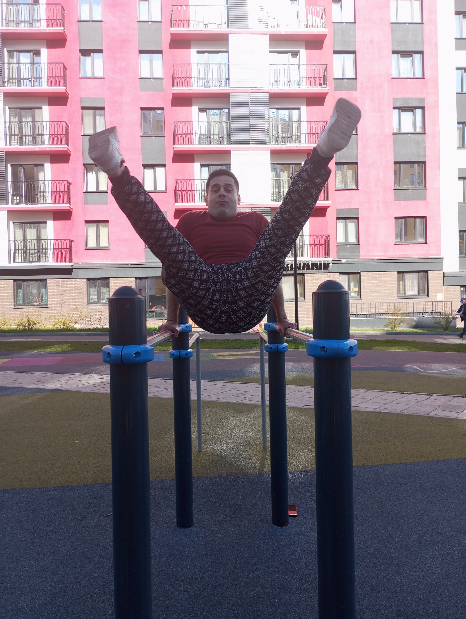 A man is performing a gymnastics routine on parallel bars in an outdoor setting. He is wearing a maroon shirt and patterned pants, with his legs spread wide and his body suspended between the bars. The parallel bars have blue fittings on top of black poles. The ground beneath the bars is a combination of gray and yellow rubberized surfaces. In the background is a tall pink building with gray accents and balconies, indicating an urban residential area. A person is walking in the far right background. The location is Pervomaysky, Russia.