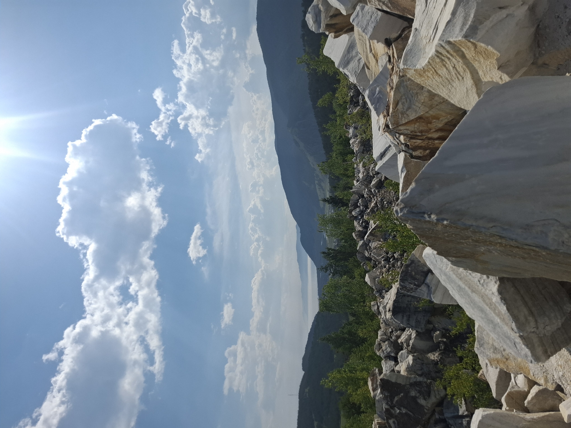 In Bugul'deika, Russia, a bright, sunny day is captured in this landscape photograph. The image features a foreground of large, white and tan marble rocks, piled haphazardly. Behind these rocks, a verdant forest stretches out, with lush green trees. In the distance, rolling hills and mountains are visible, shrouded in a slight haze. A body of water, possibly a lake or bay, separates the hills from the furthest mountains. The sky above is a vibrant blue, dotted with fluffy white clouds, and the sun shines intensely, casting a bright glare in the upper right corner. The scene depicts a natural, outdoor environment with geological formations and scenic vistas.