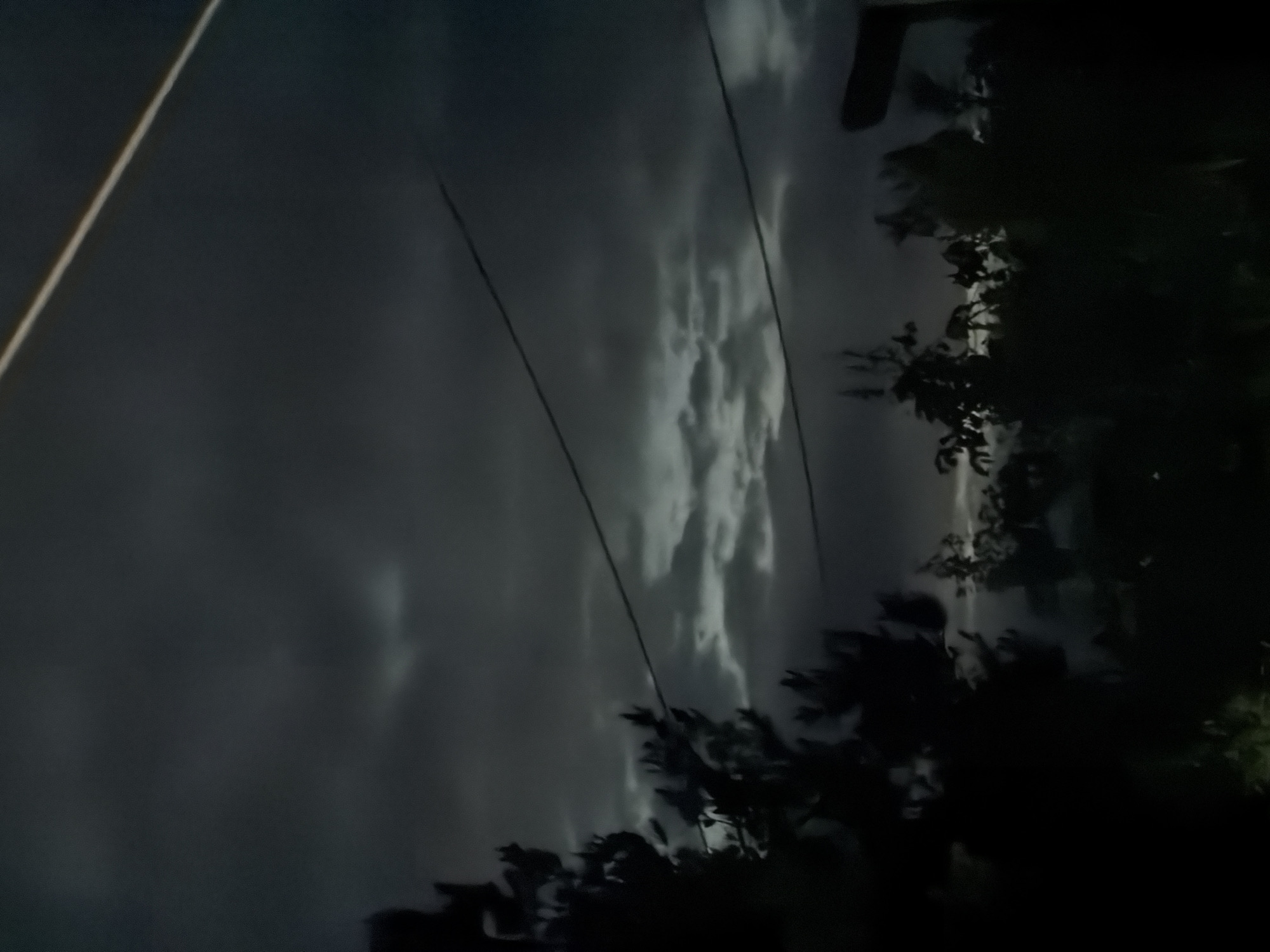 The image shows a nighttime scene, likely in a rural area, in the city of Novxanı, Azerbaijan. Dark, ominous clouds fill the sky. Two parallel wires stretch horizontally across the view. On the right side of the image, dark, indistinct shapes suggest trees and vegetation. A bright white line is present at the top left. The overall mood is somber and suggests an impending storm.