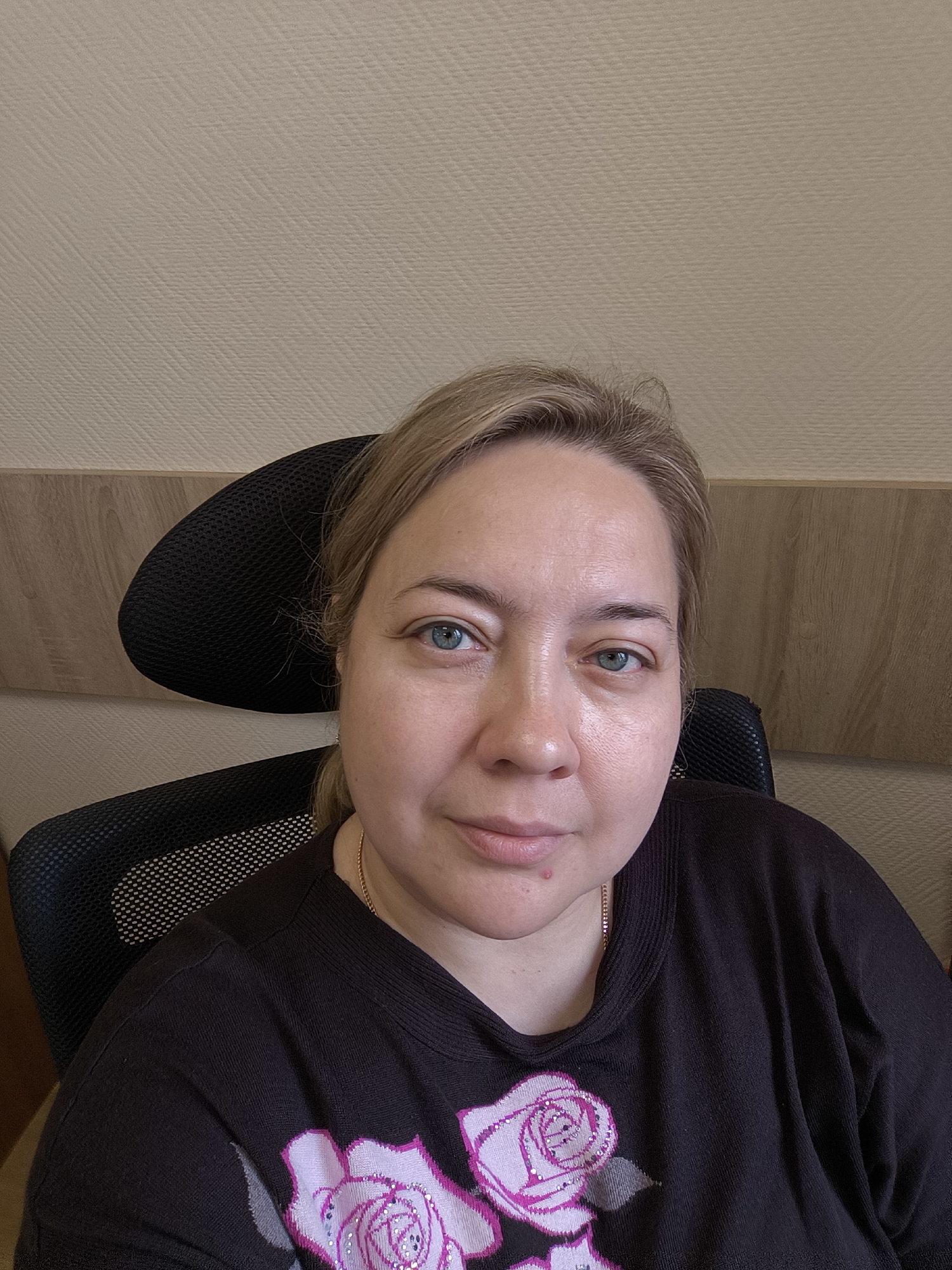 A medium close-up portrait shows a woman, likely middle-aged, looking directly at the camera. She has light skin, blue-green eyes, and blonde or light brown hair pulled back from her face. Her expression is neutral, with a slight hint of a smile. She is wearing a dark, long-sleeved top, possibly black or dark grey, featuring a large pink floral design of roses on the front, embellished with small sparkly details. A thin gold chain necklace is visible around her neck.
The woman is seated in a black office chair with a mesh backrest, parts of which are visible behind and to her side. The background consists of a light-colored, textured wall covering, possibly wallpaper, and a section of light brown wooden paneling behind the chair. This suggests an indoor setting, likely an office or home. A small red mark is visible on her chin. No specific time of day, weather conditions, or visible text are discernible in the image.