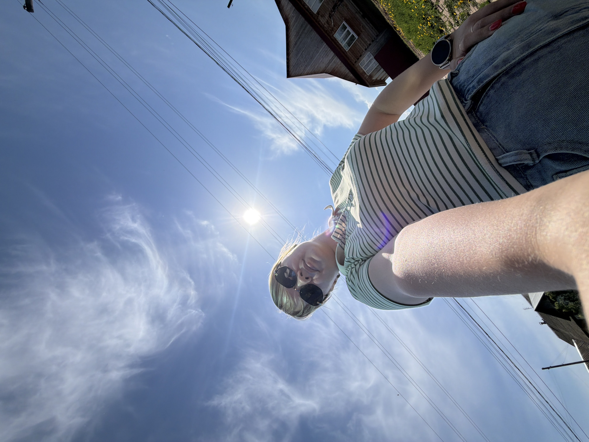 A woman is taking a selfie from a low angle. The sky is blue with wispy clouds and a bright sun. Overhead electrical wires are visible. The top of a building with yellow flowers is partially visible in the background. The woman is wearing sunglasses, a striped shirt, and jean shorts. A watch is on her wrist. The location is Чумна, Russia.