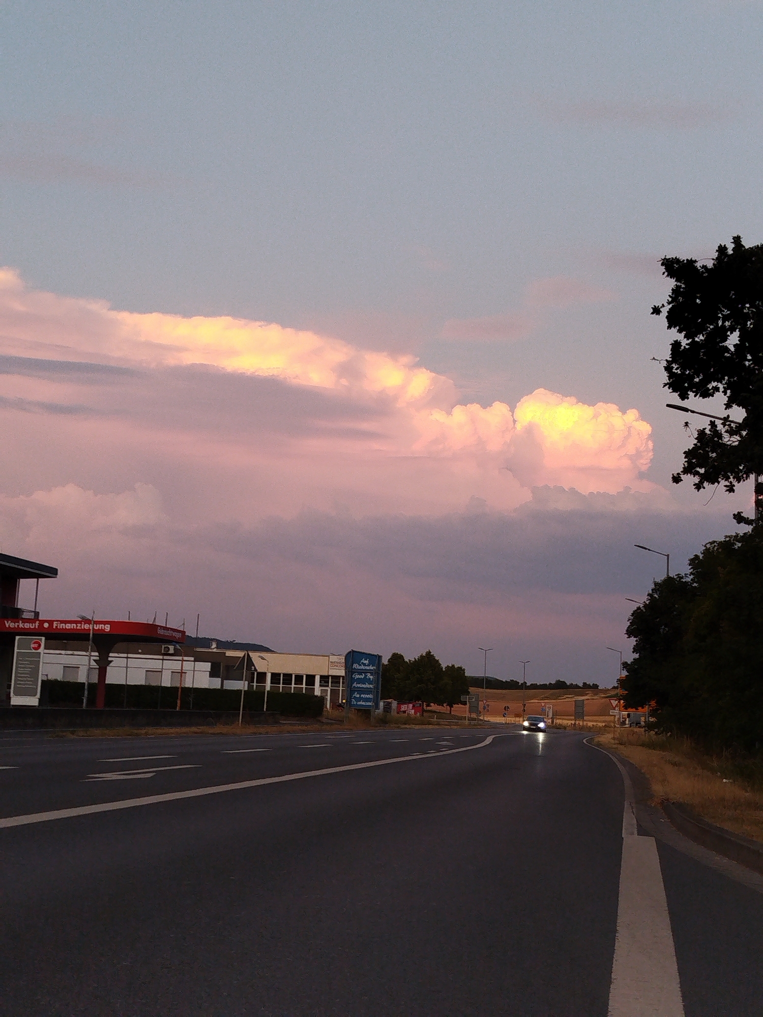 The image depicts a wide, paved road in the city of Kitzingen, Germany, at dusk. The sky is filled with soft, pastel-colored clouds, tinged with pink and yellow from the setting sun. The road curves gently to the right, with white lane markings visible. A dark-colored car with its headlights on is driving away from the viewer in the distance, on the right side of the road.
Along the left side of the road, there is a building with a red awning that reads "Verkauf • Finanzierung" (Sales • Financing) in white lettering. Adjacent to this building is a modern-looking structure with a white facade and dark-framed windows. Several streetlights are visible, lining the road on both sides, and casting long shadows. On the right side of the road, lush green trees and vegetation line the embankment. In the background, there are more buildings and structures, some with visible signage. The overall atmosphere is serene and tranquil, characteristic of a twilight scene.
