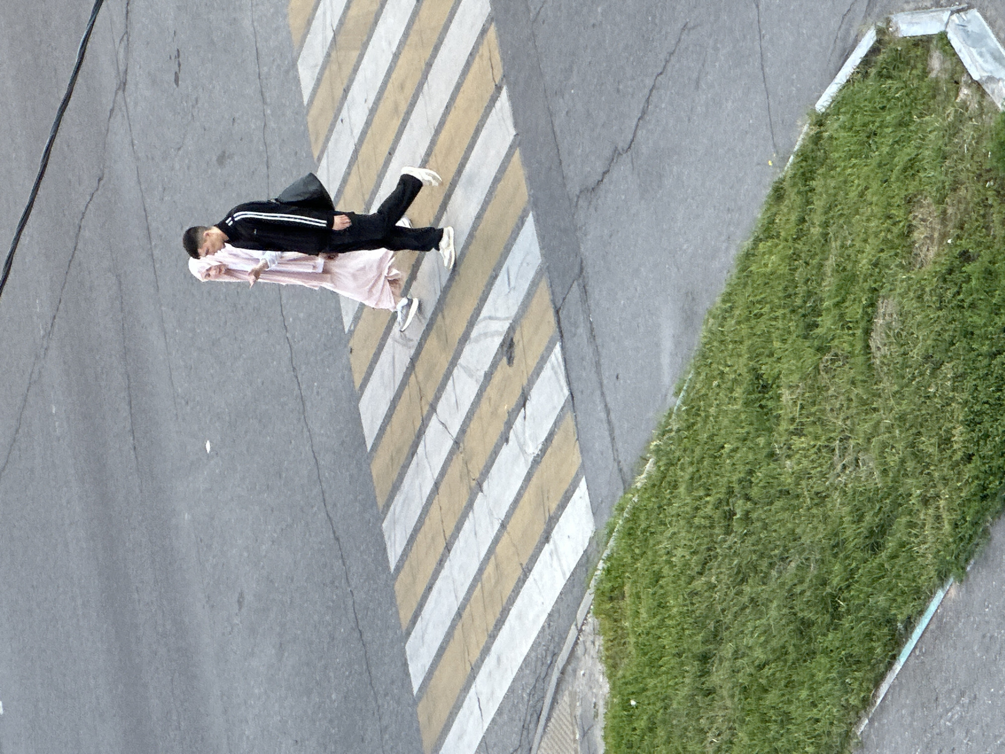 Two individuals are crossing a crosswalk in Podolsk, Russia. On the left, a person in a light pink hijab and a long dress is walking. To their right, a person wearing a black tracksuit with white stripes down the sleeves and pants, and carrying a dark bag over their shoulder, is walking. The crosswalk has alternating yellow and white stripes. The ground is asphalt, and there is a patch of green grass to the lower left of the frame, bordered by a concrete curb. The scene is viewed from an elevated perspective, looking down on the street.