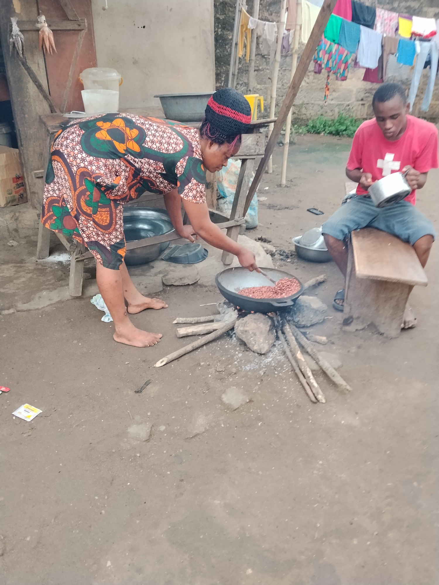 In an outdoor setting in an unspecified city and country, an adult female wearing a patterned dress and a knitted cap is bent over, stirring the contents of a large, dark-colored pan. The pan rests on a fire built on the ground using small pieces of wood and rocks. Nearby, an adult male in a red t-shirt and blue shorts is sitting on a small, wooden stool, holding a small metal pot. Laundry hangs on a clothesline in the background.