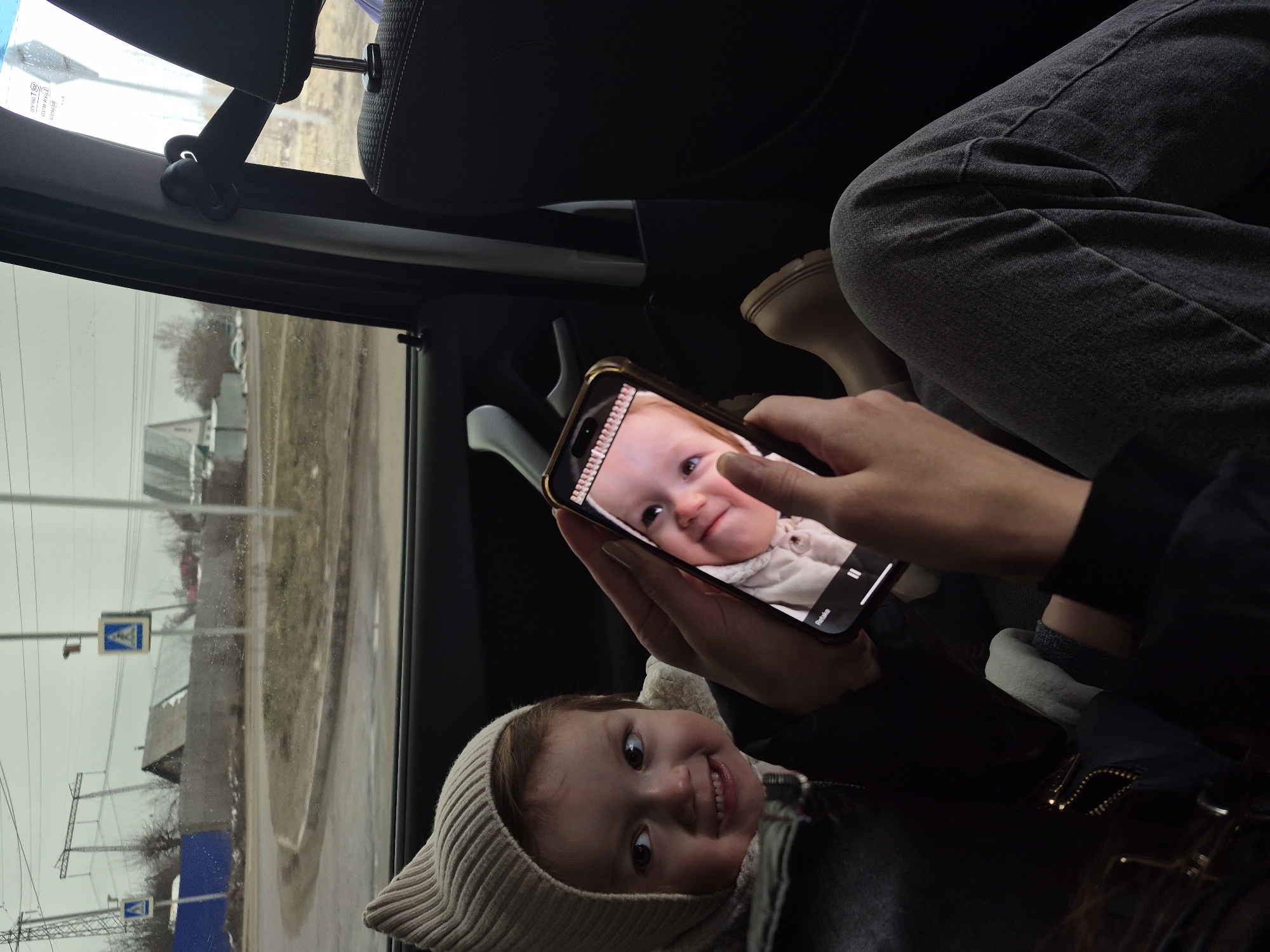 A medium shot, taken from within a car, shows a young girl wearing a light-colored, cat-eared hat, smiling directly at the camera. The girl is in the lower portion of the frame. A hand is holding a mobile phone with a photo of a baby with similar features, slightly turned towards the left. To the upper left of the frame, the exterior is visible through the car window, with a pedestrian crossing sign and buildings suggesting an urban or suburban area. The shot is taken in Chaikovsky, Russia, as evidenced by the pedestrian sign, which is specific to that country. The lighting suggests an overcast day.