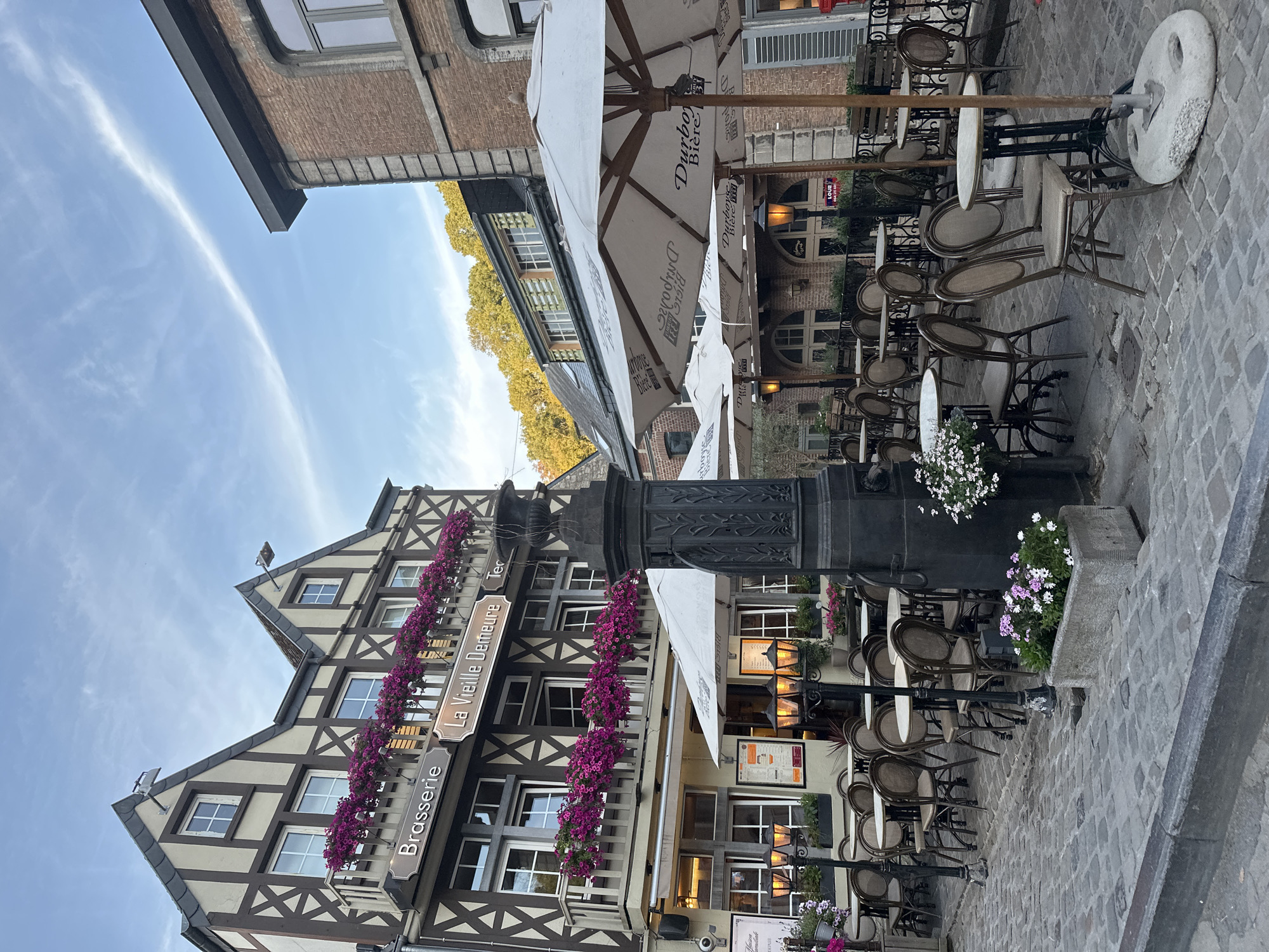 The image depicts an outdoor seating area of a brasserie in Durbuy, Belgium. The brasserie, named "La Vieille Demeure," is housed in a half-timbered building with a balcony adorned with purple flowers. There are several tables and chairs set up on a cobblestone street, some covered by large white umbrellas. Streetlights illuminate the area, and there are potted plants and a stone fountain in the foreground. The sky is a pale blue with some wispy clouds. The scene appears to be quiet, with no people visible. The overall atmosphere is one of charm and relaxation, suggesting a pleasant place to dine outdoors.