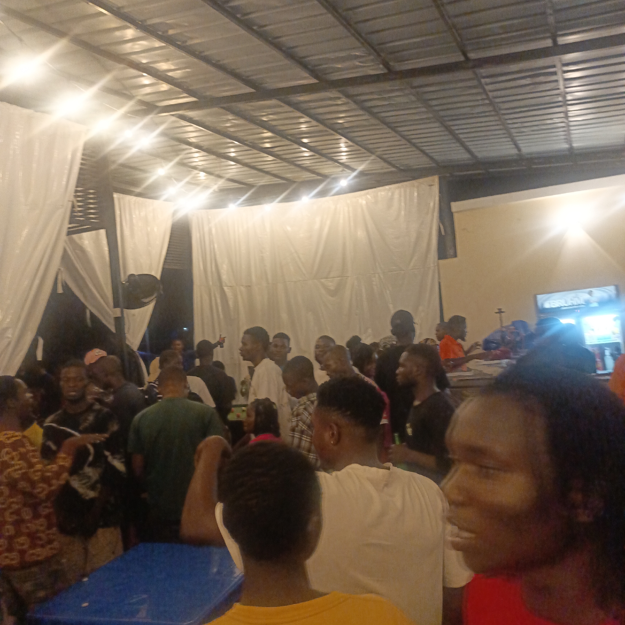 A medium shot shows a gathering of people in an indoor space. The location is presumed to be in Ube, Nigeria. The space has a metal roof structure with visible supports and lights. White drapery is hung along the walls.
Many individuals are present, their clothing varying in style and color. Some are standing, while others appear to be seated at tables. Several people seem to be interacting, and some are gesturing with their hands.
A television screen is visible in the background. A blue table is in the foreground.