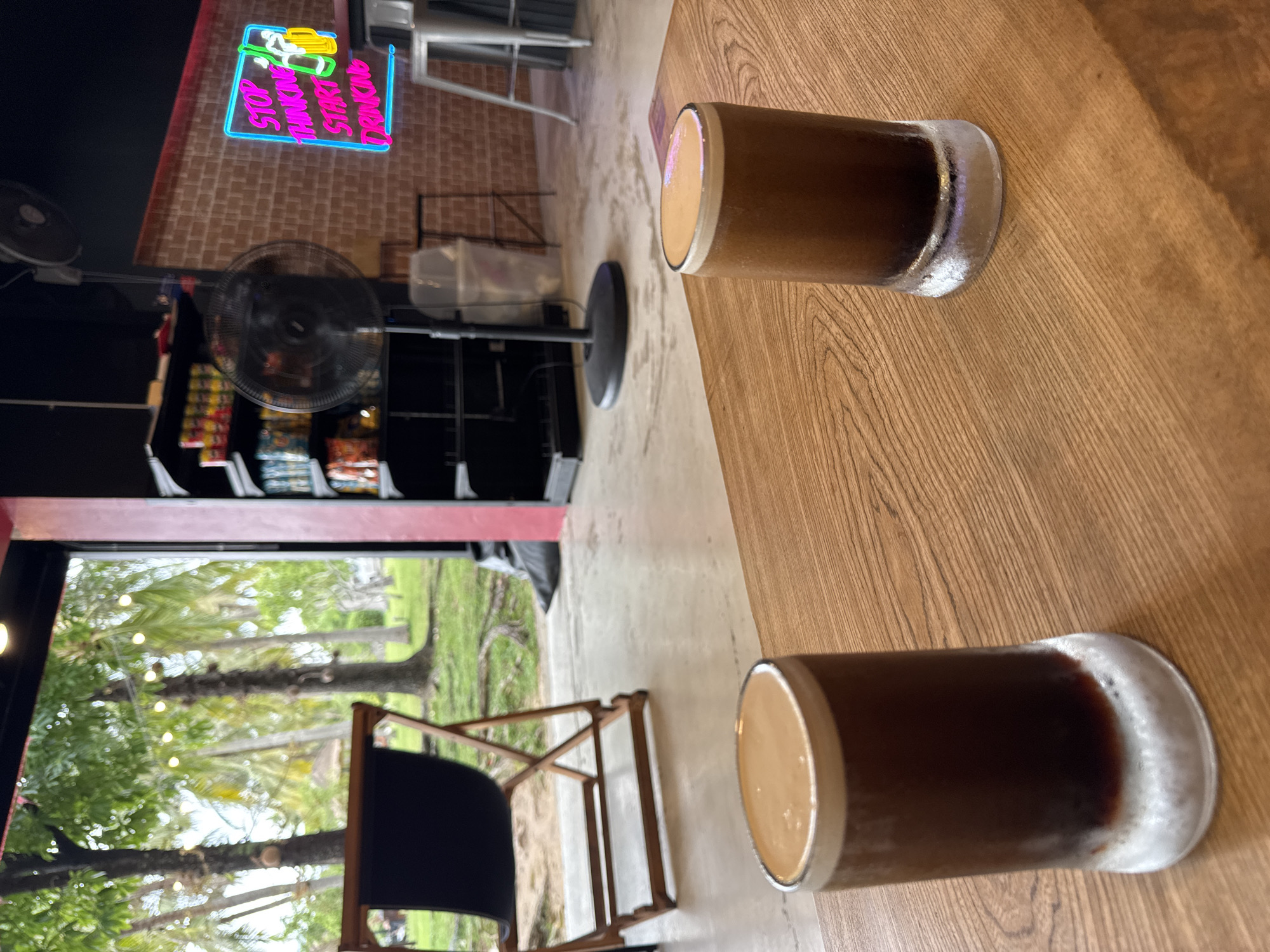 The image shows two glasses of dark-colored beverages with a creamy top, likely coffee or beer, placed on a wooden table in what appears to be an establishment in บ้านบางรักษ์, Thailand. Outside, visible through a window, are trees and foliage, possibly a garden or park. Inside, a folding chair is partially visible and a floor fan. A neon sign with the words "STOP THINKING START DRINKING" is mounted on a brick wall. A rack of snacks is visible in the background.