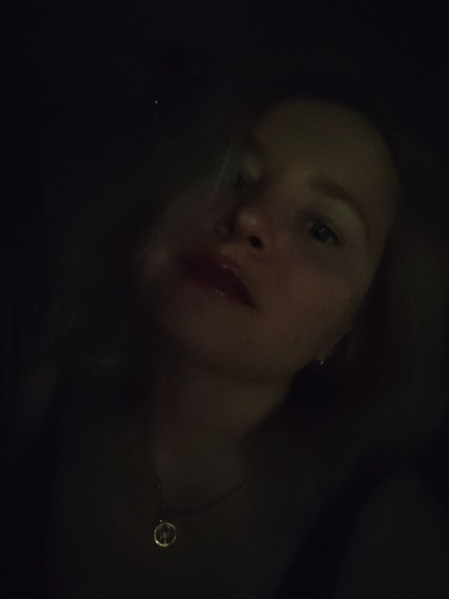 The image features a close-up portrait of a young woman in an extremely dimly lit environment, likely at night or indoors with minimal lighting. Her face, partially illuminated, is positioned diagonally, with her head slightly tilted. She has light-colored hair, which is visible around her face. Her eyes are open and she appears to be looking towards the camera. Her lips are distinctly visible due to a bright red lipstick. A dark, possibly metallic, round pendant hangs from a necklace around her neck, reflecting some light. The background is almost entirely dark, offering no discernible details of the setting beyond the implied low-light conditions. No specific location cues are visible within the image, nor is there any visible text. The overall mood is intimate and subdued due to the low lighting.