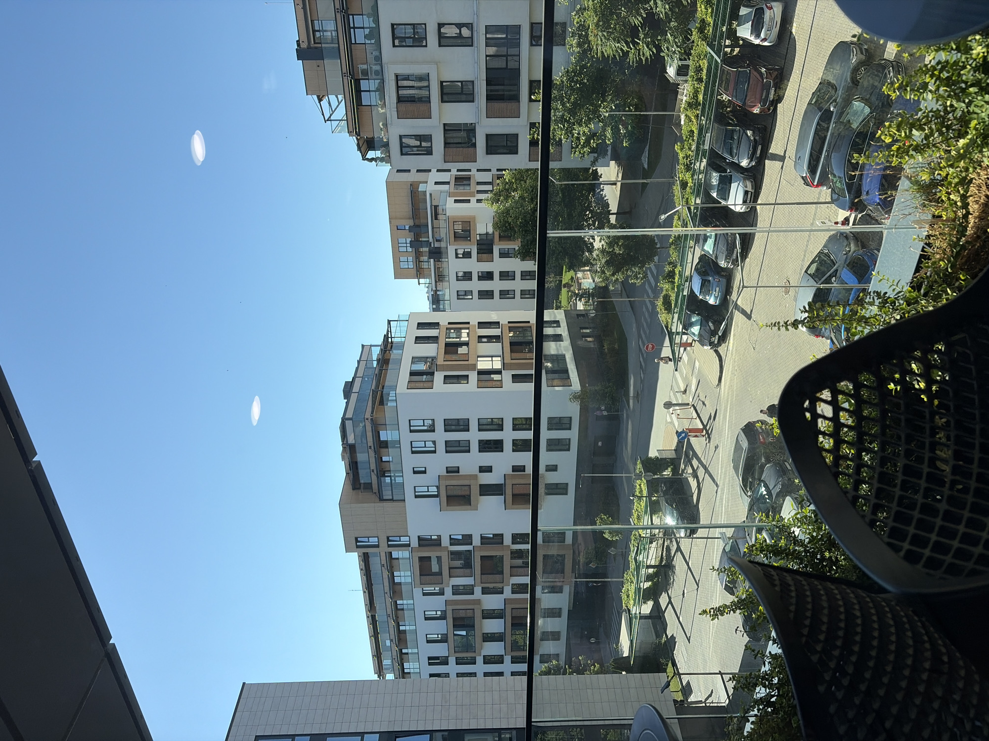 In a city environment, modern apartment buildings with balconies and white facades are visible under a clear blue sky. In the foreground, two dark, intricate outdoor chairs are positioned in front of a glass railing, overlooking a street. Cars are parked in a lot in front of the buildings, and trees line the street and parking area. One circular, bright object is visible in the sky.