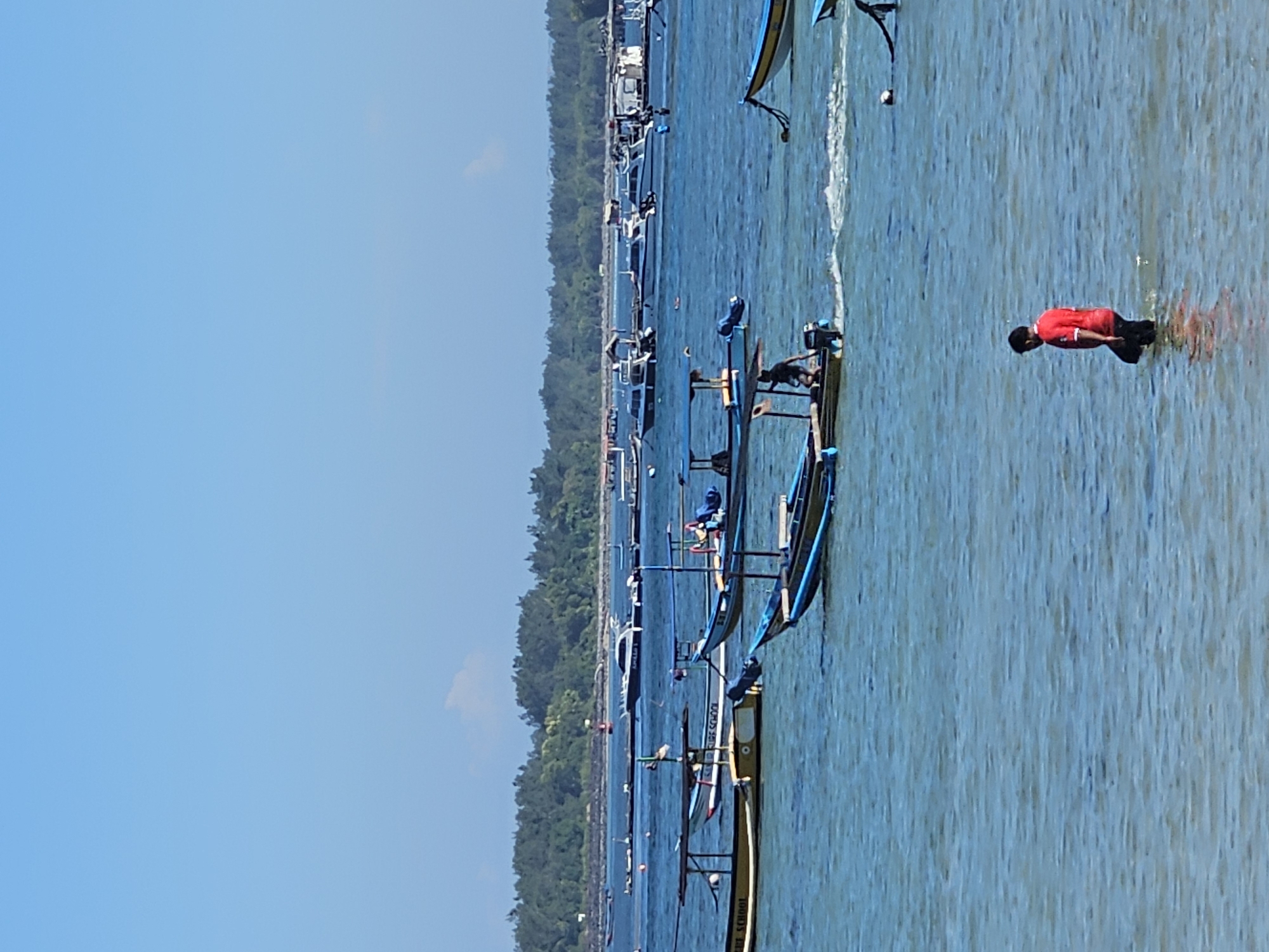 A bright, clear day unfolds over a coastal scene in Denpasar, Indonesia. The foreground is dominated by the shimmering, shallow blue waters of the ocean, where a lone man in a red shirt and black pants wades, his legs submerged. In the mid-ground, a collection of traditional Indonesian fishing boats, known as jukungs, bob on the gentle waves. Several of these boats are equipped with outboard motors. One jukung is actively moving, leaving a wake, with a person at the helm. Other boats are anchored or drifting. Beyond the boats, a lush green treeline forms the distant horizon, meeting a pale blue sky dotted with a few wispy clouds. The overall impression is one of serene coastal activity.