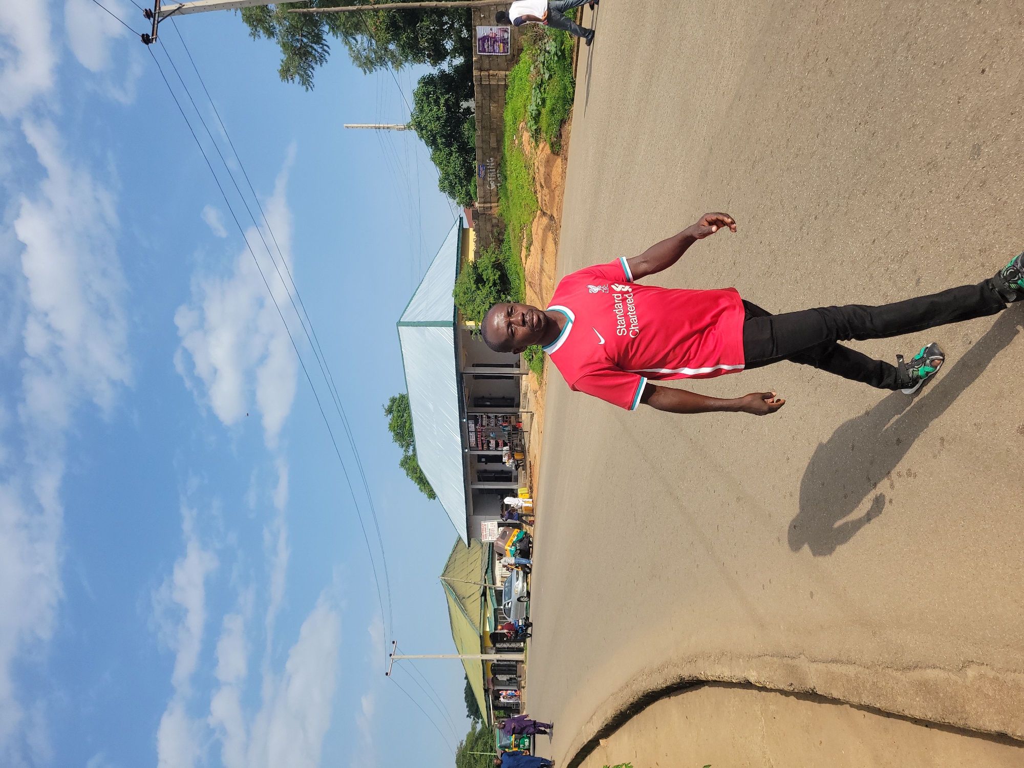 A medium shot shows a Black man walking on an asphalt road, his body angled slightly to the right. He wears a red short-sleeved t-shirt with white trim and a Liverpool F.C. crest, black pants, and green and black sandals. His shadow is cast on the road beside him. In the background, to the left, a brown embankment lines the road. Further back are buildings with light-colored walls and green or silver roofs, some with awnings. A few people and vehicles, including a motorcycle and a trishaw, are visible in front of the buildings. Trees and a telephone pole with wires are also present. The sky above is blue with scattered white clouds.