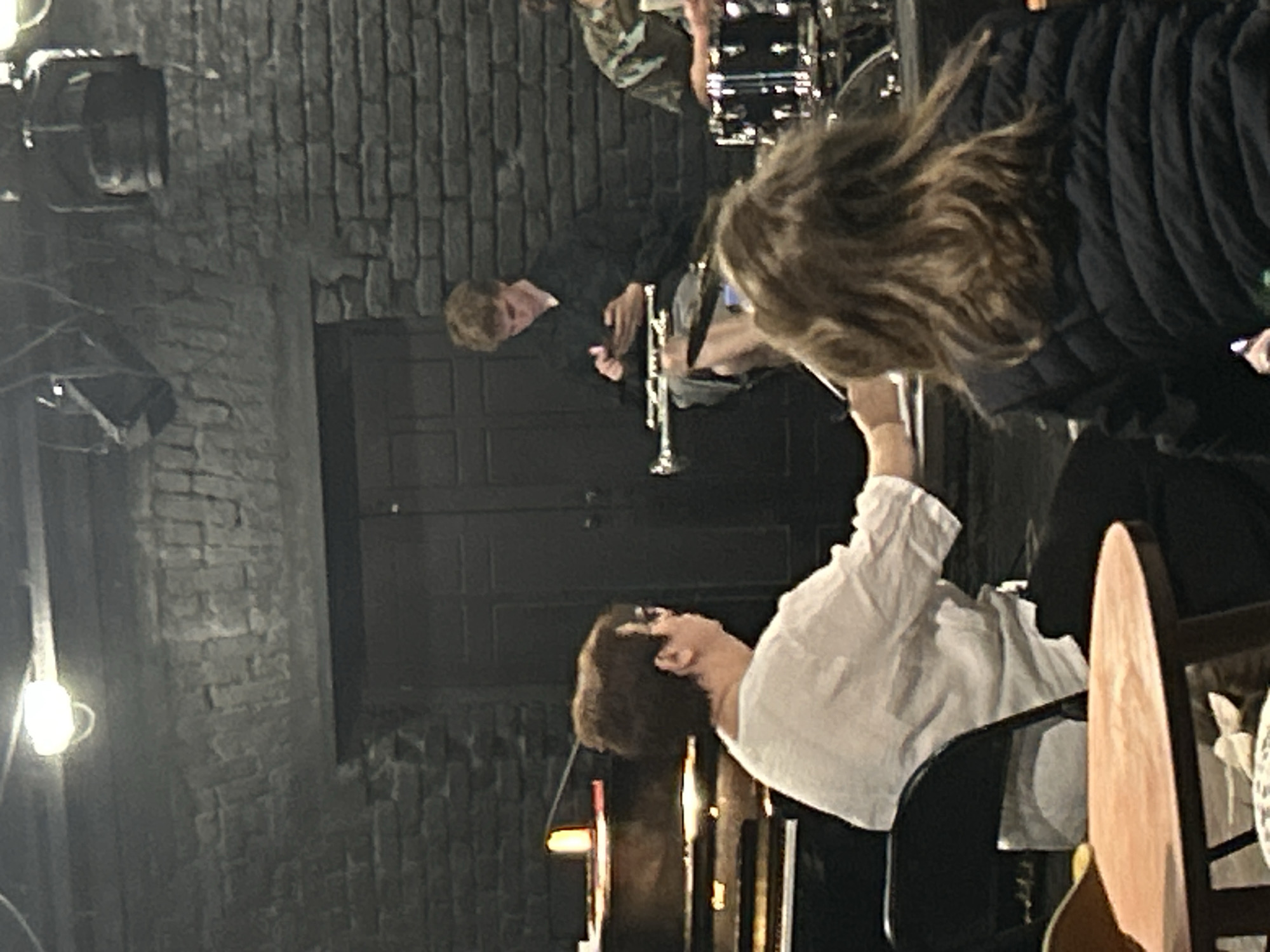 The image shows a dimly lit, indoor scene with exposed brick walls. A musician plays a trumpet in front of a closed, dark-colored double door. Another musician is behind him playing drums. In the foreground, a person wearing a white shirt is seated, facing the stage, and a person with long hair wearing a black puffer jacket is standing in the right foreground. Several chairs are also visible. The location is Unknown, Unknown.