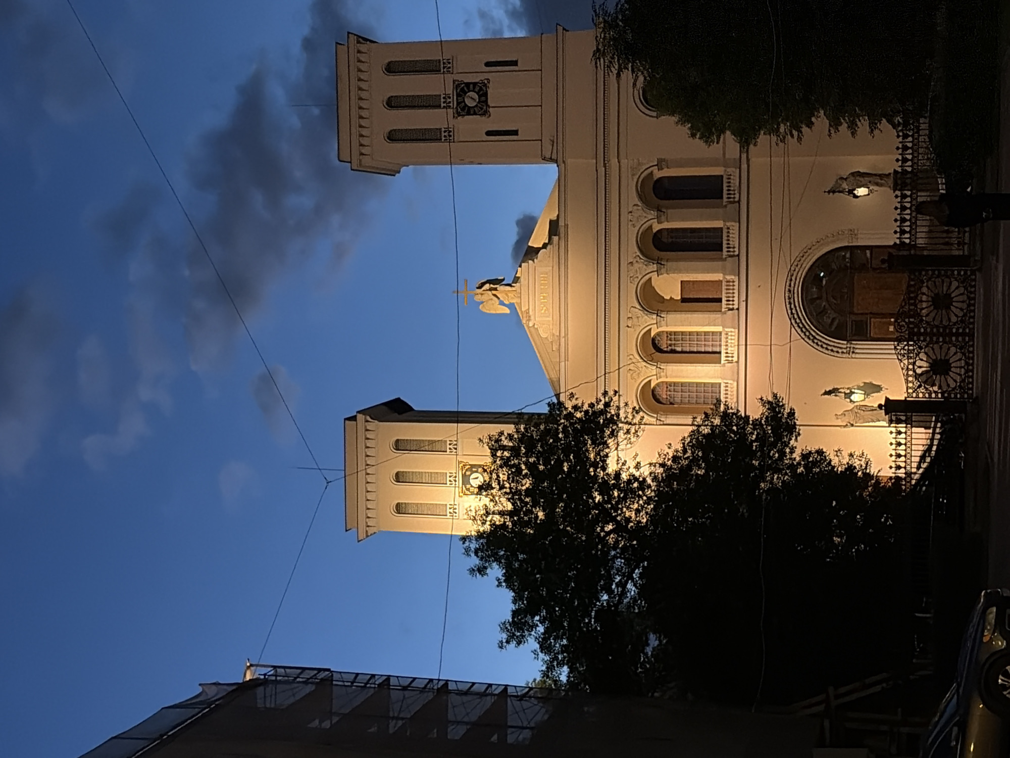 The image depicts the Lutheran Church of Saint Peter and Saint Paul in Saint Petersburg, Russia, at dusk. The church features two prominent bell towers, one with a visible clock face displaying the time as approximately 4:10. The facade of the church is illuminated, highlighting architectural details such as arched windows and a central entrance with ornate gates. A statue of an angel with a cross stands on the roof above the entrance. Silhouetted trees are visible in the foreground, partially obscuring the lower portion of the church. The sky is a deep blue with scattered clouds, indicating the transition from day to night. A dark-colored car is parked on the left side of the frame, and a person in dark clothing is standing near the church gates on the right. Electrical wires crisscross the sky above the building. The overall scene captures a serene, atmospheric view of the historic church at the end of the day.