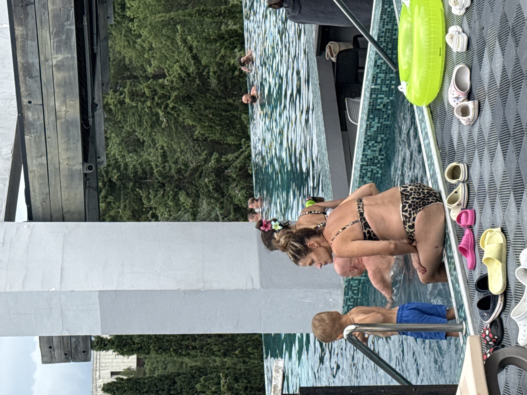 The image shows a group of people in and around a swimming pool on a bright, overcast day. Several individuals are swimming in the clear blue water, with some enjoying the refreshing dip while others appear to be relaxing. A woman in a leopard-print bikini is leaning forward, looking into the pool, while a man in blue swim trunks stands nearby. A young boy is also present, looking towards the water.
The pool area is surrounded by lush green trees, suggesting a natural or well-landscaped setting. The poolside features a textured grey surface with several pairs of colorful flip-flops and clogs neatly arranged, including pink, yellow, and white options. A bright green inflatable ring floats in the water, indicating recreational use of the pool.
In the background, a wooden structure is visible, possibly a deck or part of a building. The overall atmosphere appears casual and relaxed, with people enjoying a day at the pool. There are no visible signs of text in the image. The setting suggests a warm weather destination, likely a resort or private pool area.