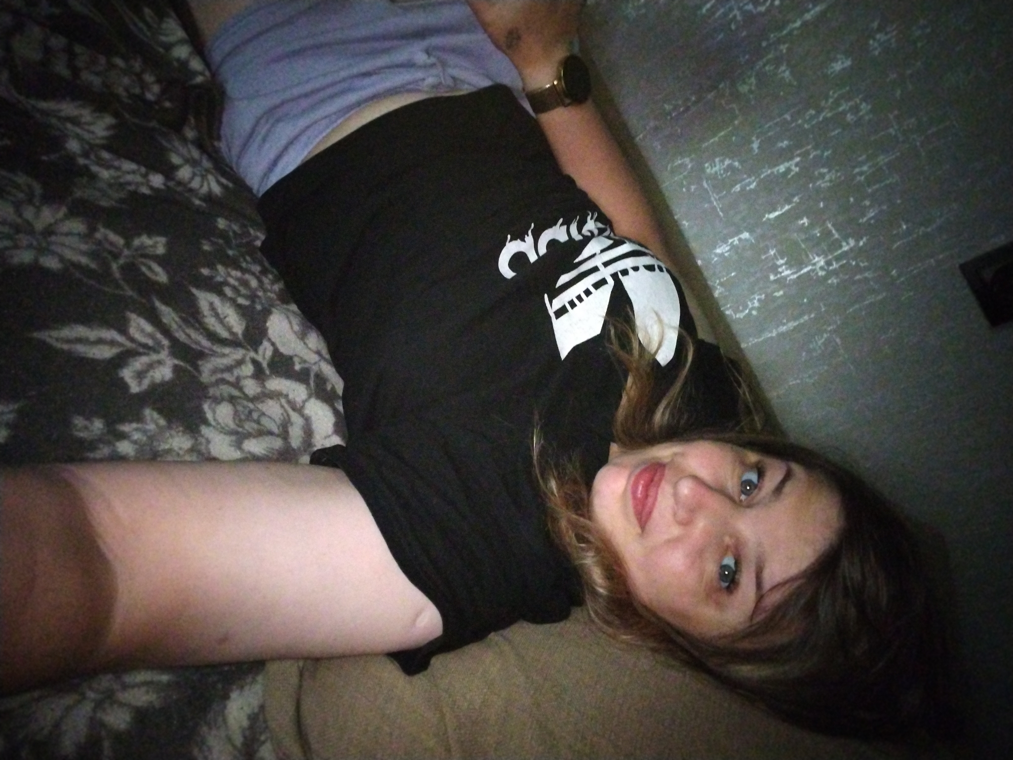 The image depicts a young woman lying on a bed. She is wearing a black t-shirt with a white graphic design and a purple garment. The room appears to be dimly lit, with a textured wall in the background. There is a floral patterned blanket on the bed. The woman is looking up at the camera with her head tilted. Her right arm is visible, resting on her leg, and she is wearing a watch on her left wrist. The image does not contain enough information to determine the location within the city of Yaroslavl, Russia.