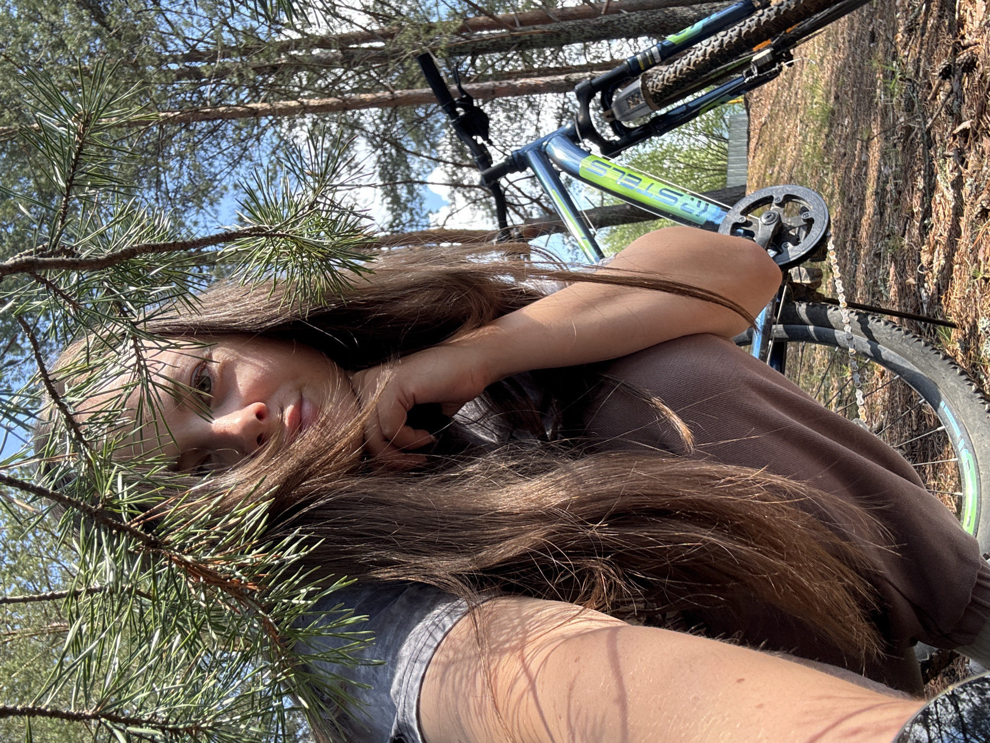 The image shows a young woman with long, brown hair reclining next to a bicycle in a wooded area. The woman is wearing a brown sweatshirt and has her head resting on her hand. A branch with pine needles is partially obscuring her face. The bicycle is positioned to the right of the woman. The location appears to be outdoors, with trees and a clear sky visible in the background. There is no information given in the picture that indicates that it was taken in Малая Рукавицкая, Russia.