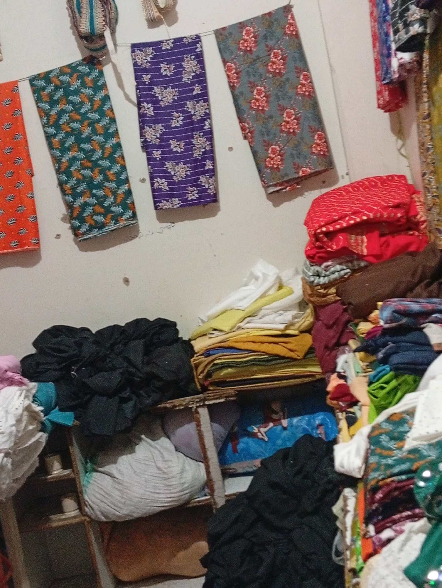 The image depicts a cluttered storage area, likely a textile shop, in Larkana, Pakistan. Hanging on a plain wall are three folded pieces of patterned fabric: one orange with a white leaf-like pattern, one green with a similar pattern, and one purple with white stripes and floral motifs. To the right, a stack of folded red fabric with a textured pattern is visible, along with other folded fabrics in various colors and textures. Below the hanging fabrics, shelves are filled with piles of fabric and clothing. One shelf contains a large bundle of white fabric. Another shelf holds a large black pile of fabric, and a blue item with a pattern is partially visible. The overall impression is one of an abundant but disorganized textile inventory. The lighting is dim and appears to be artificial.